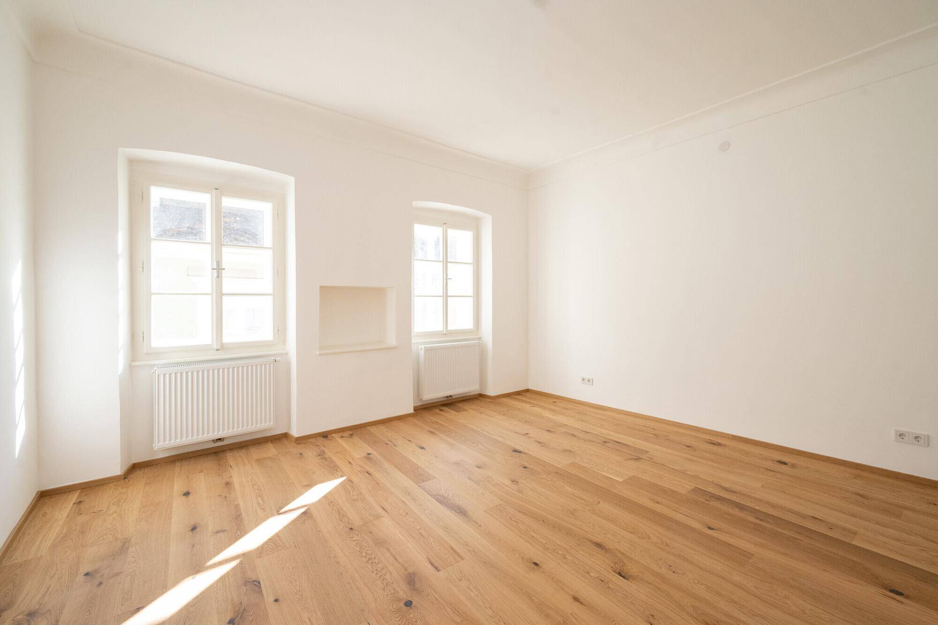 Helles Zimmer mit Holzboden und Heizkörper unter den Fenstern, frisch renoviert.
