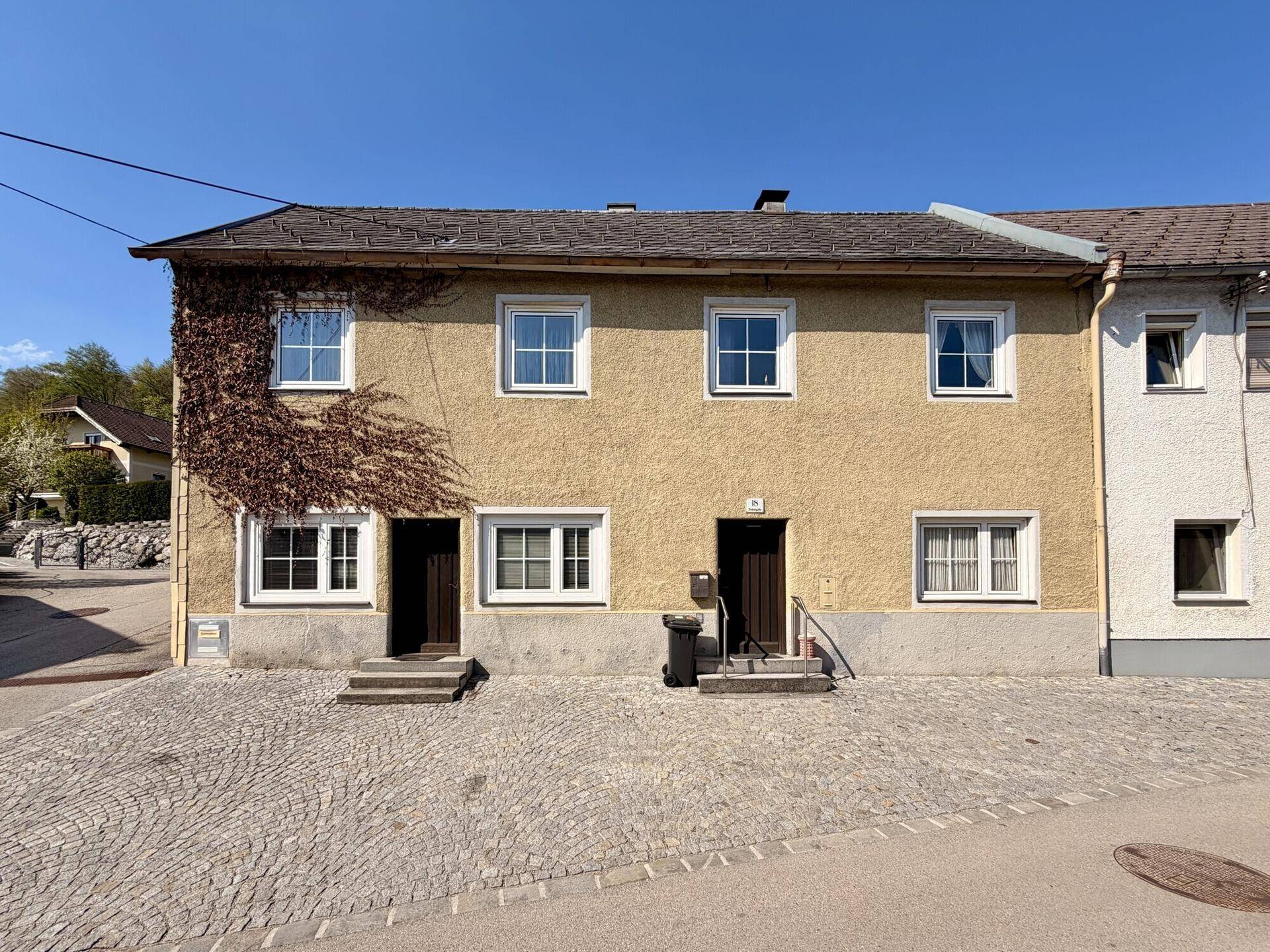Gelbes Reihenhaus mit bewachsener Fassade und gepflastertem Vorplatz unter blauem Himmel.