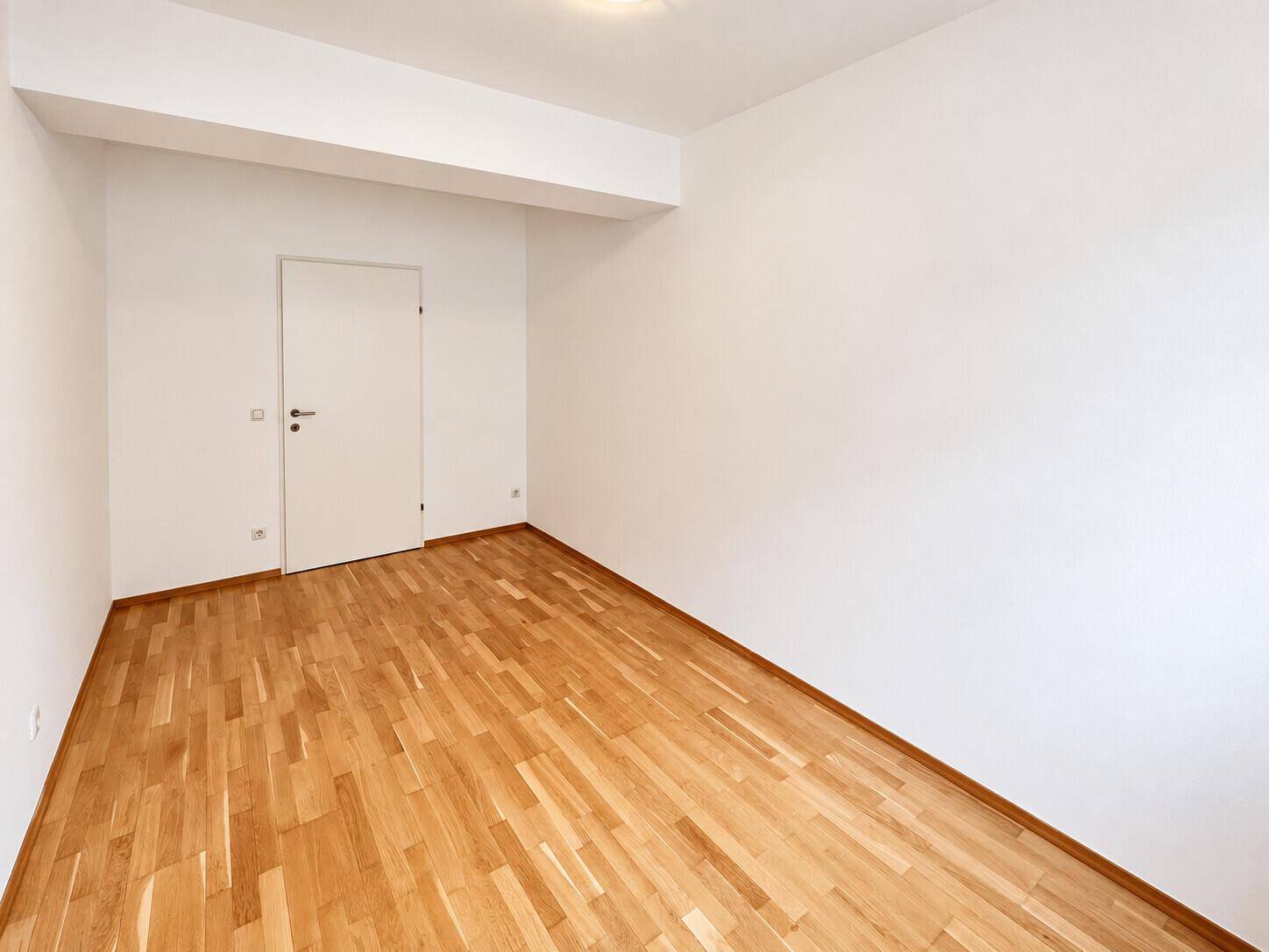 Leeres Zimmer mit Parkettboden und weißer Tür, bietet flexible Nutzungsmöglichkeiten für Bewohner.