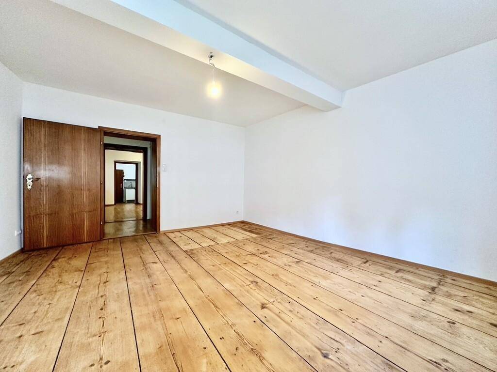 Leerer Raum mit Holzdielenboden und Zugang zu weiteren Zimmern durch eine Holztür.