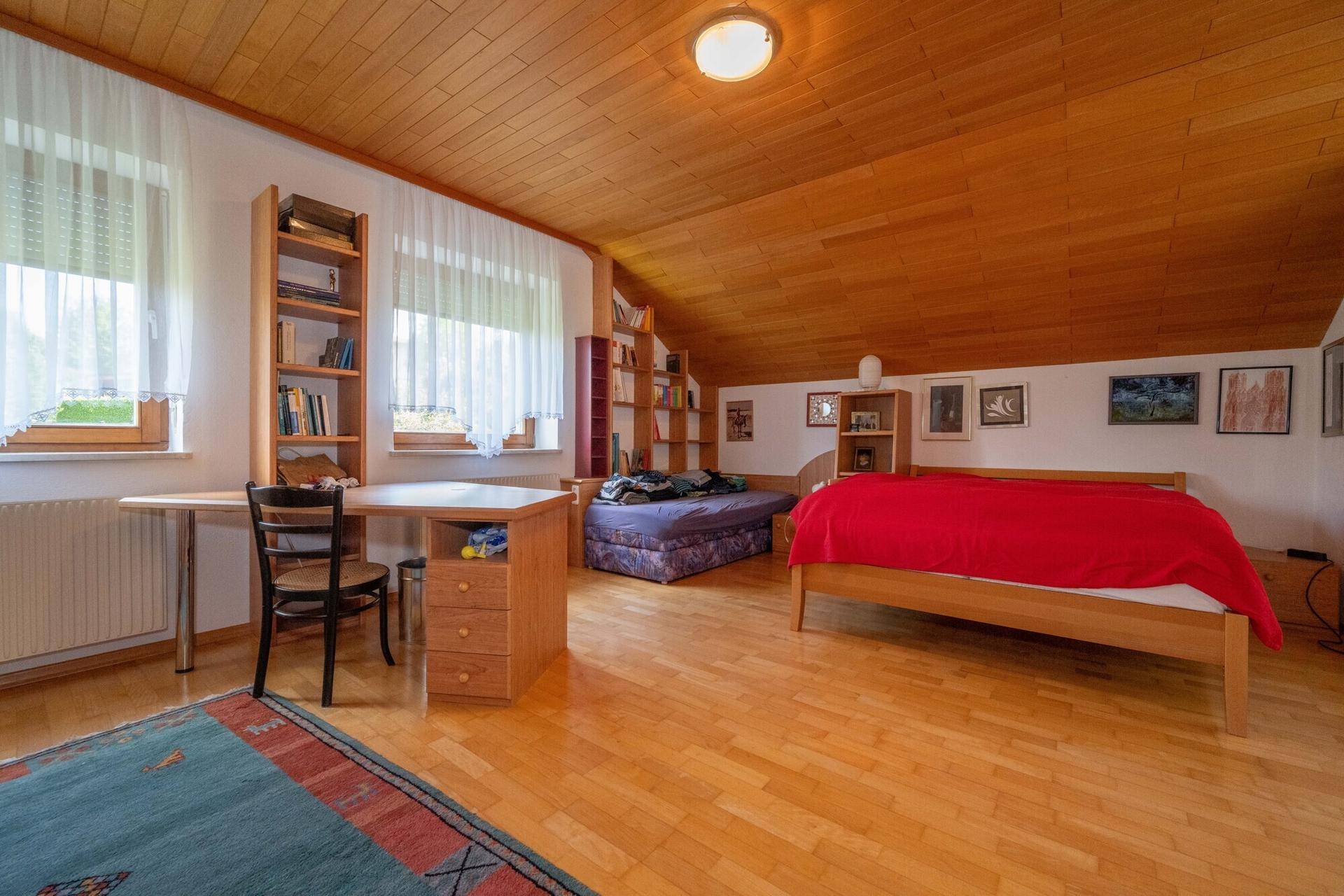Schlafzimmer mit Holzdecke, Bett und Schreibtischbereich unter dem Dachschrägen.