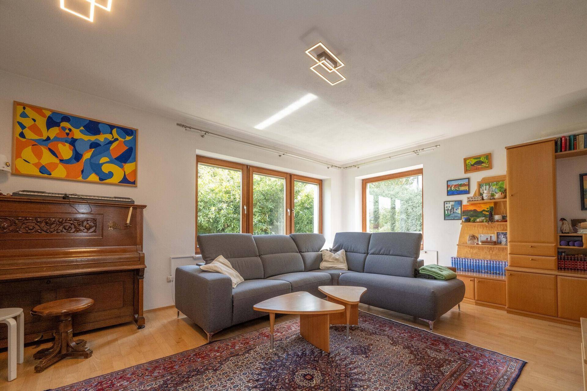 Geräumiges Wohnzimmer mit bequemer Couch, Klavier und großen Fenstern mit Blick ins Grüne.