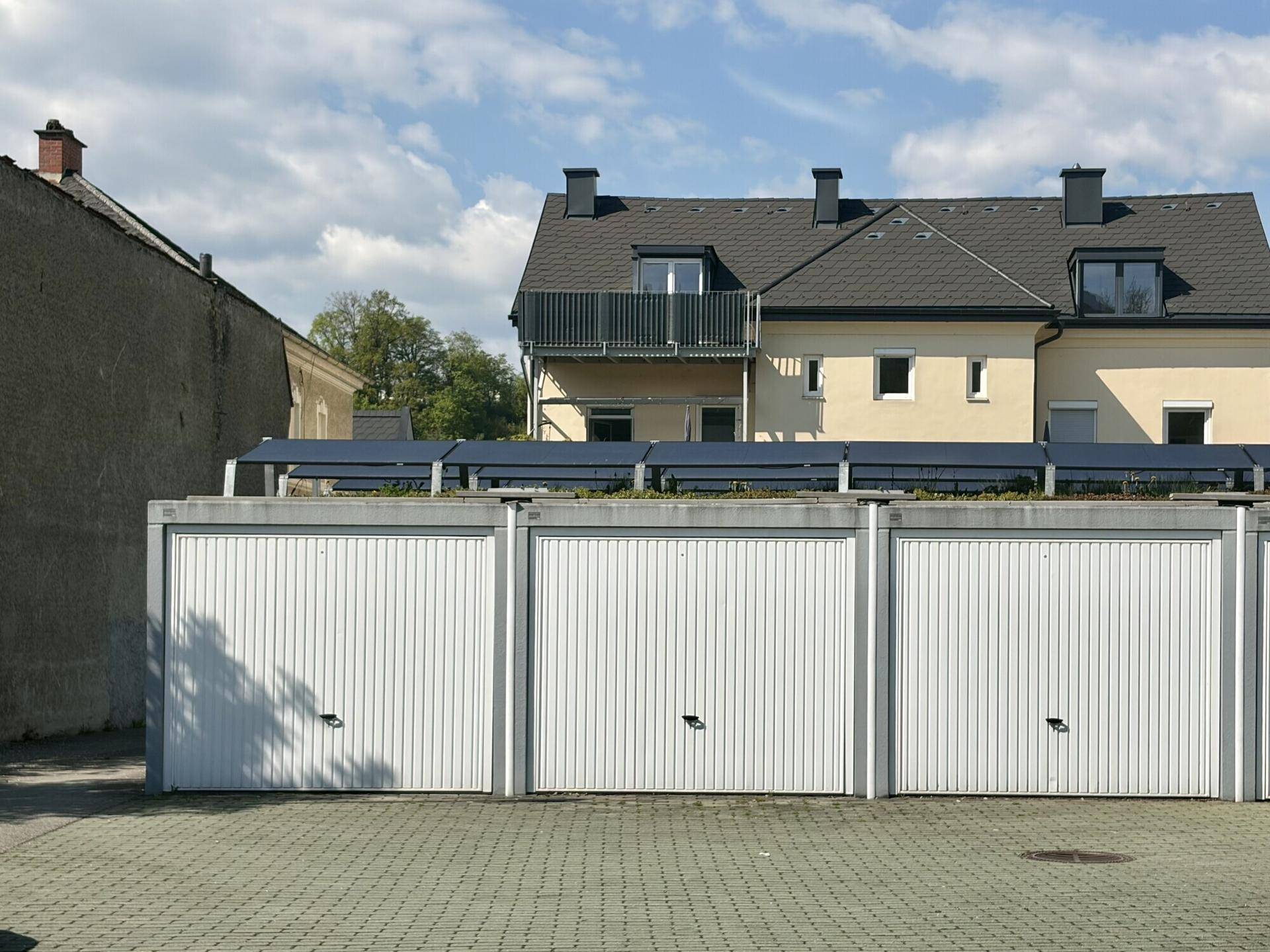 Drei Garagenboxen mit weißen Toren vor einem Wohngebäude mit Balkon unter blauem Himmel.