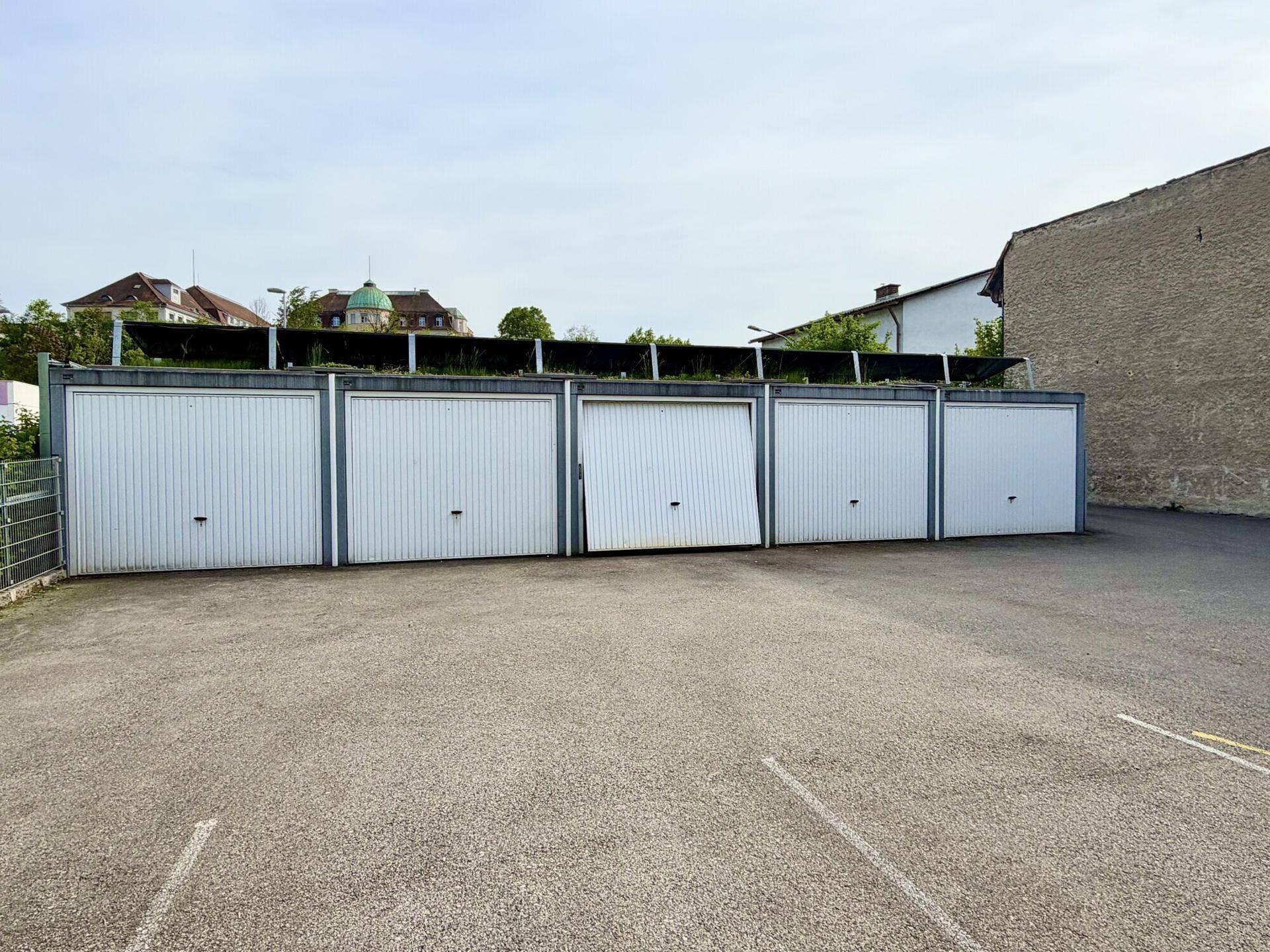 Reihe von Garagenboxen mit weißen Toren und einem gepflasterten Vorplatz unter hellem Himmel.