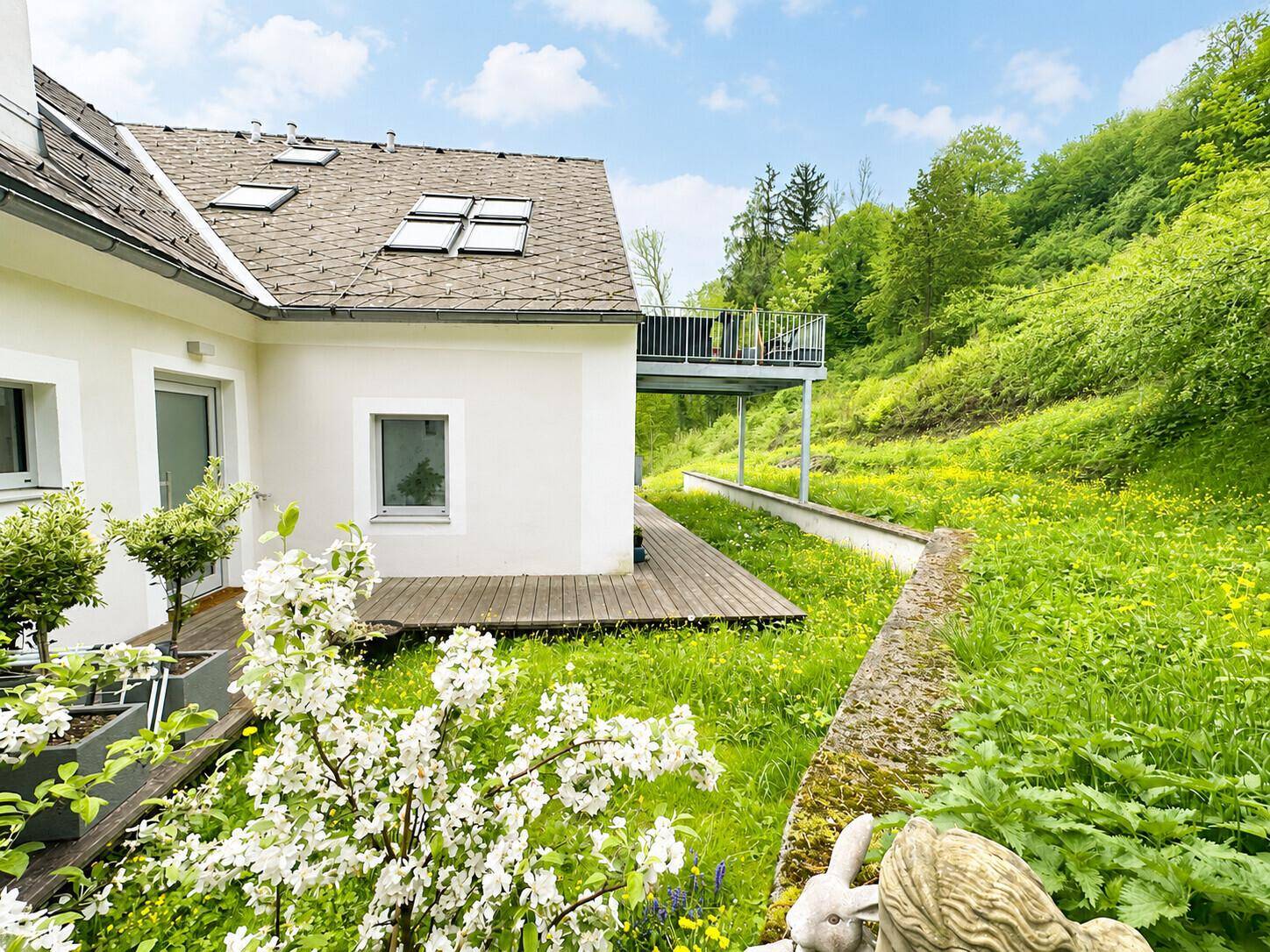 Hausfassade mit Balkon und Holzterrasse, umgeben von grünen Hügeln und Natur.