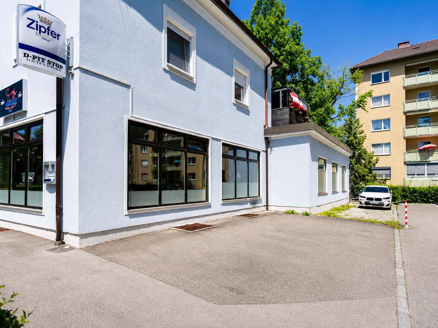 Nahaufnahme der blauen Gebäudefassade mit großen Fenstern und einem kleinen Parkplatz davor.