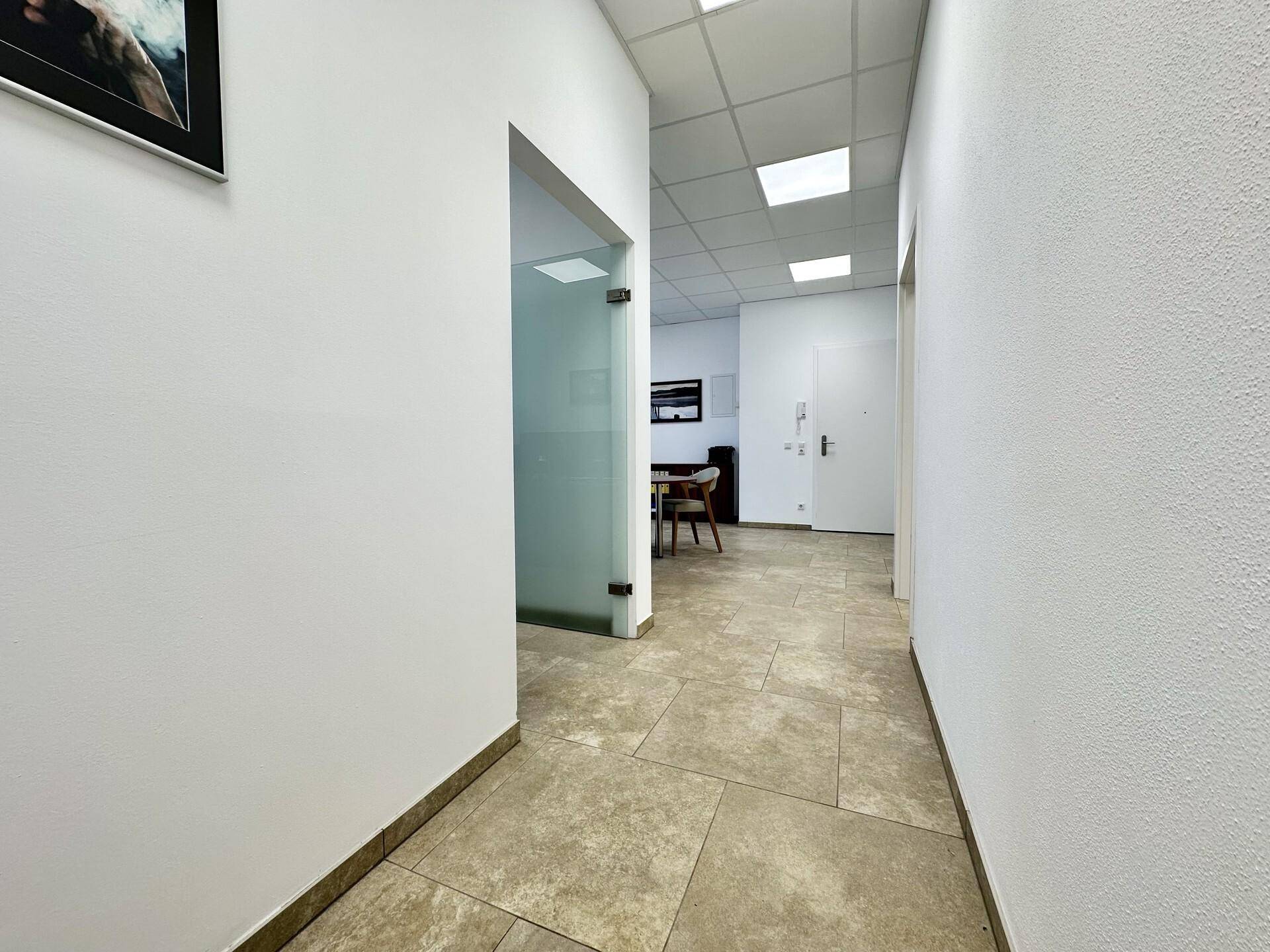 Heller Flur mit modernem Fliesenboden, weißen Wänden und einer Glastür, die zu einem Büro führt.