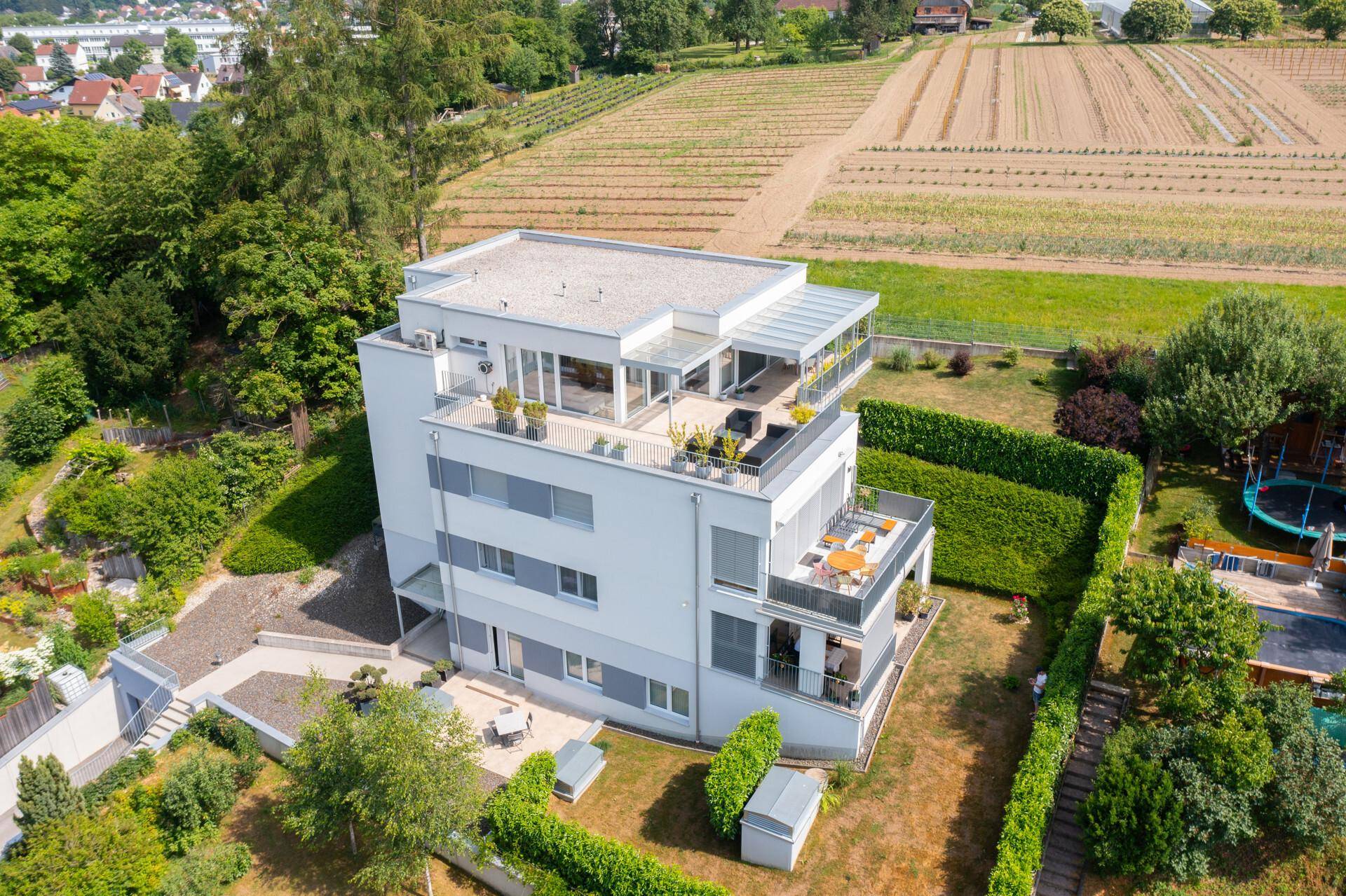 Luftaufnahme eines modernen Mehrfamilienhauses mit mehreren Terrassen und Balkonen, umgeben von grünen Feldern.