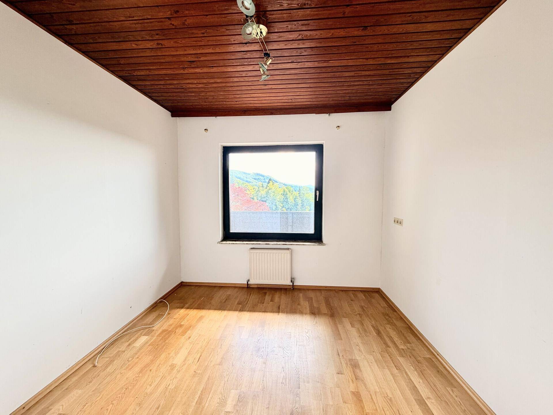 Leeres Zimmer mit Holzdecke, Laminatboden und Fenster mit Ausblick.