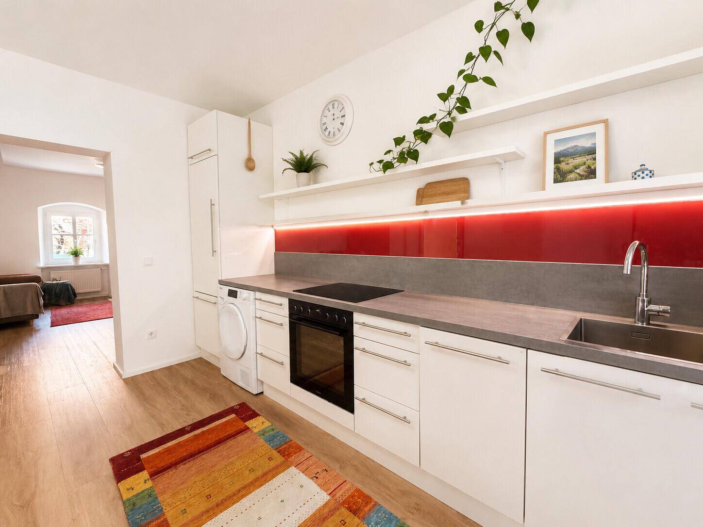 Moderne Einbauküche mit weißer Front, rotem Spritzschutz und Blick in den angrenzenden Wohnbereich.