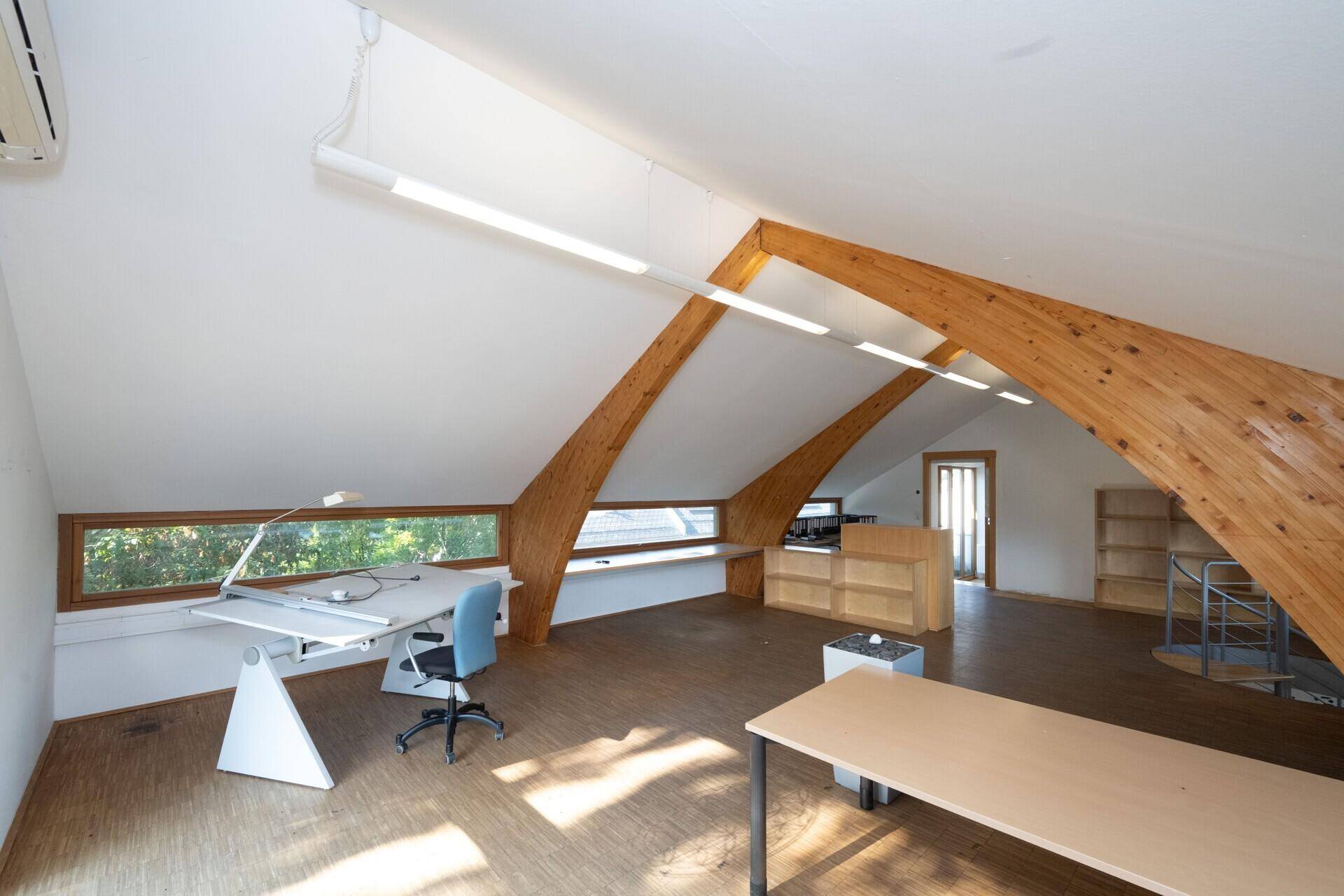 Dachgeschossbüro mit Klimaanlage, Holzbalken und Arbeitsplätzen unter den Fenstern.