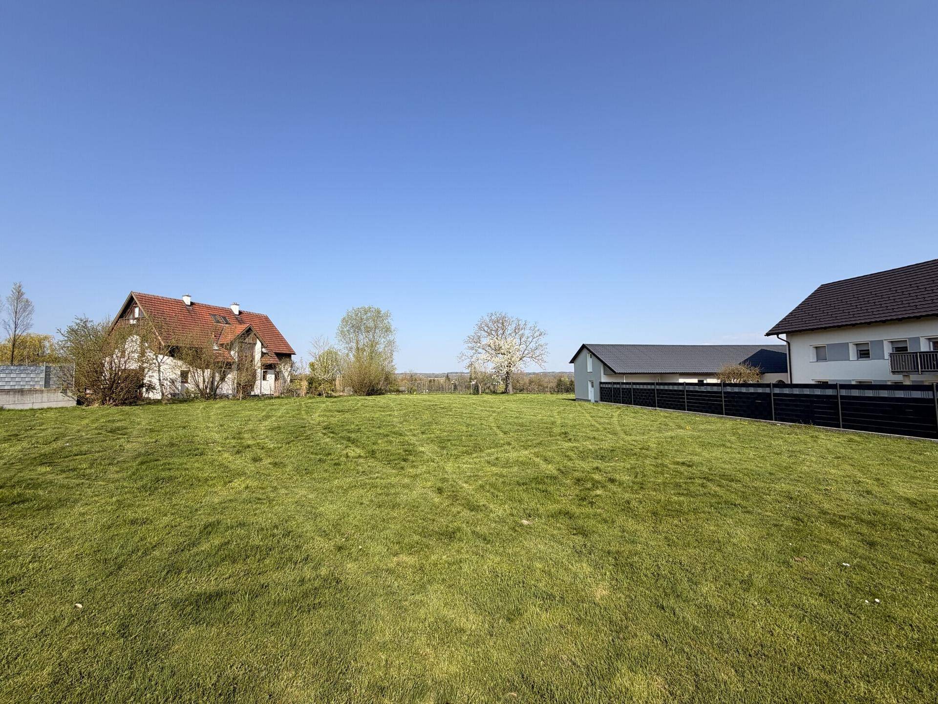 Weitläufige grüne Wiese mit traditionellen und modernen Häusern im Hintergrund und Fernsicht.