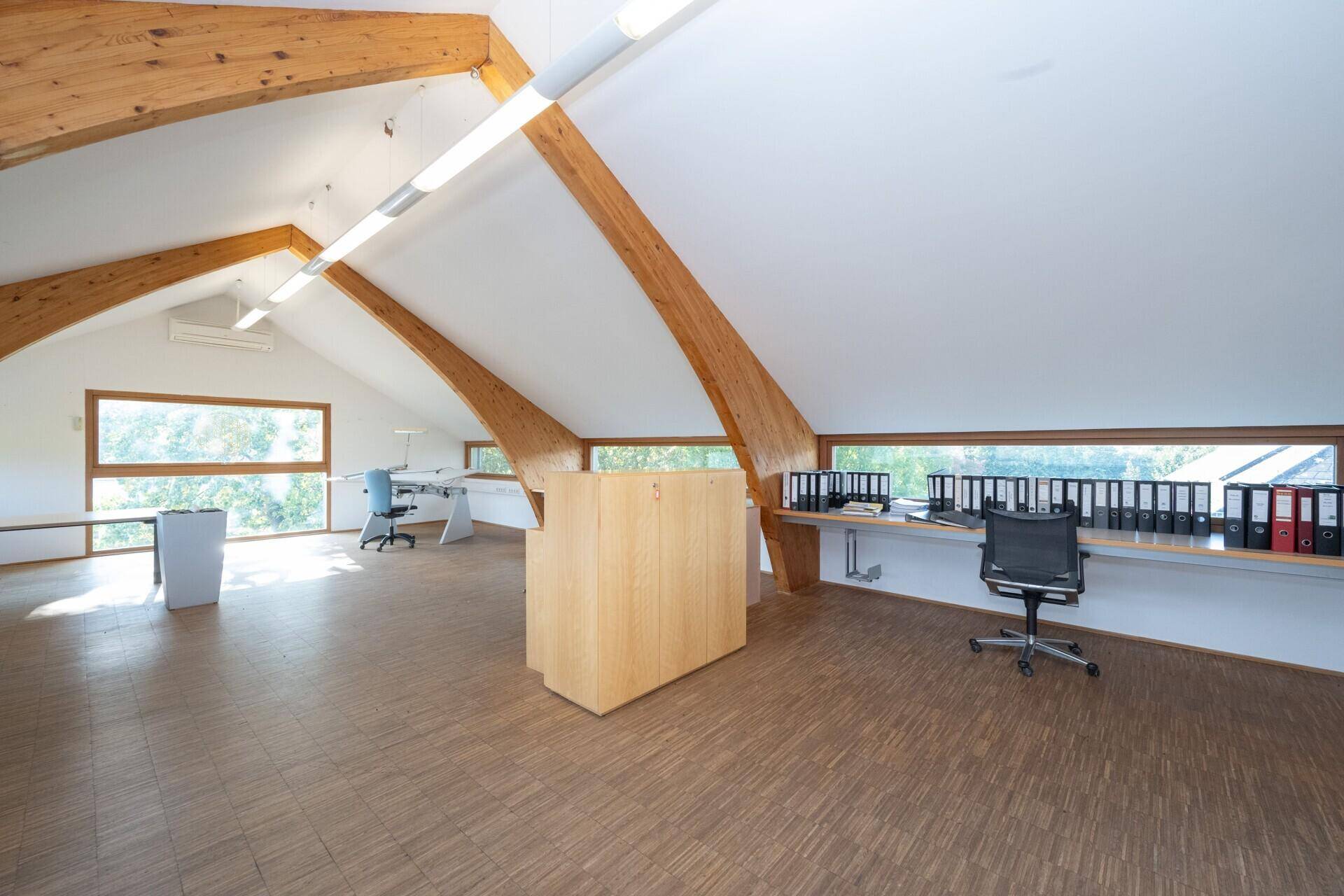 Geräumiges Dachgeschossbüro mit sichtbaren Holzbalken, schrägen Decken und großen Fenstern.