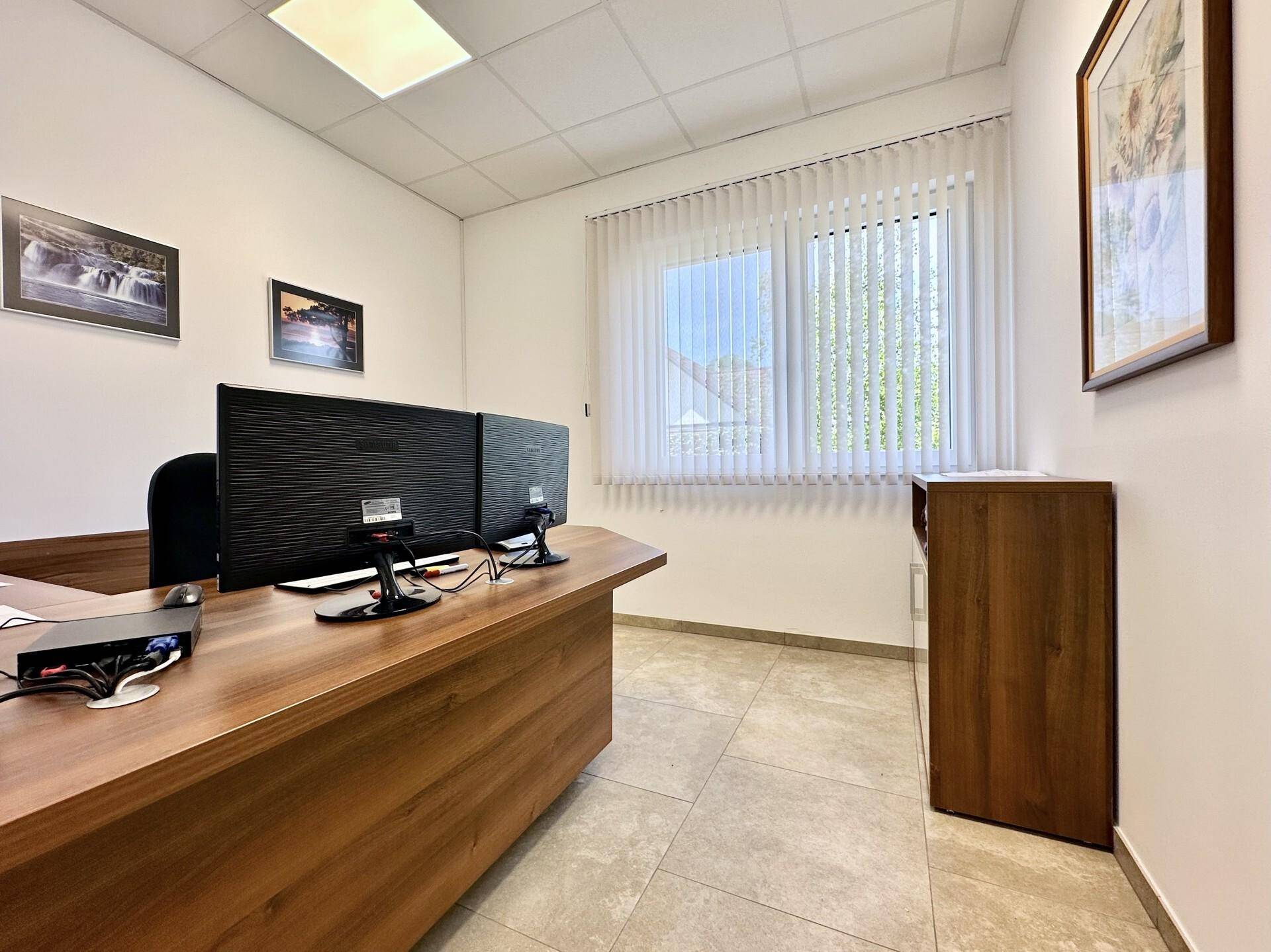 Helles Büro mit einem Holzschreibtisch, Computermonitoren und einem Fenster mit vertikalen Jalousien.