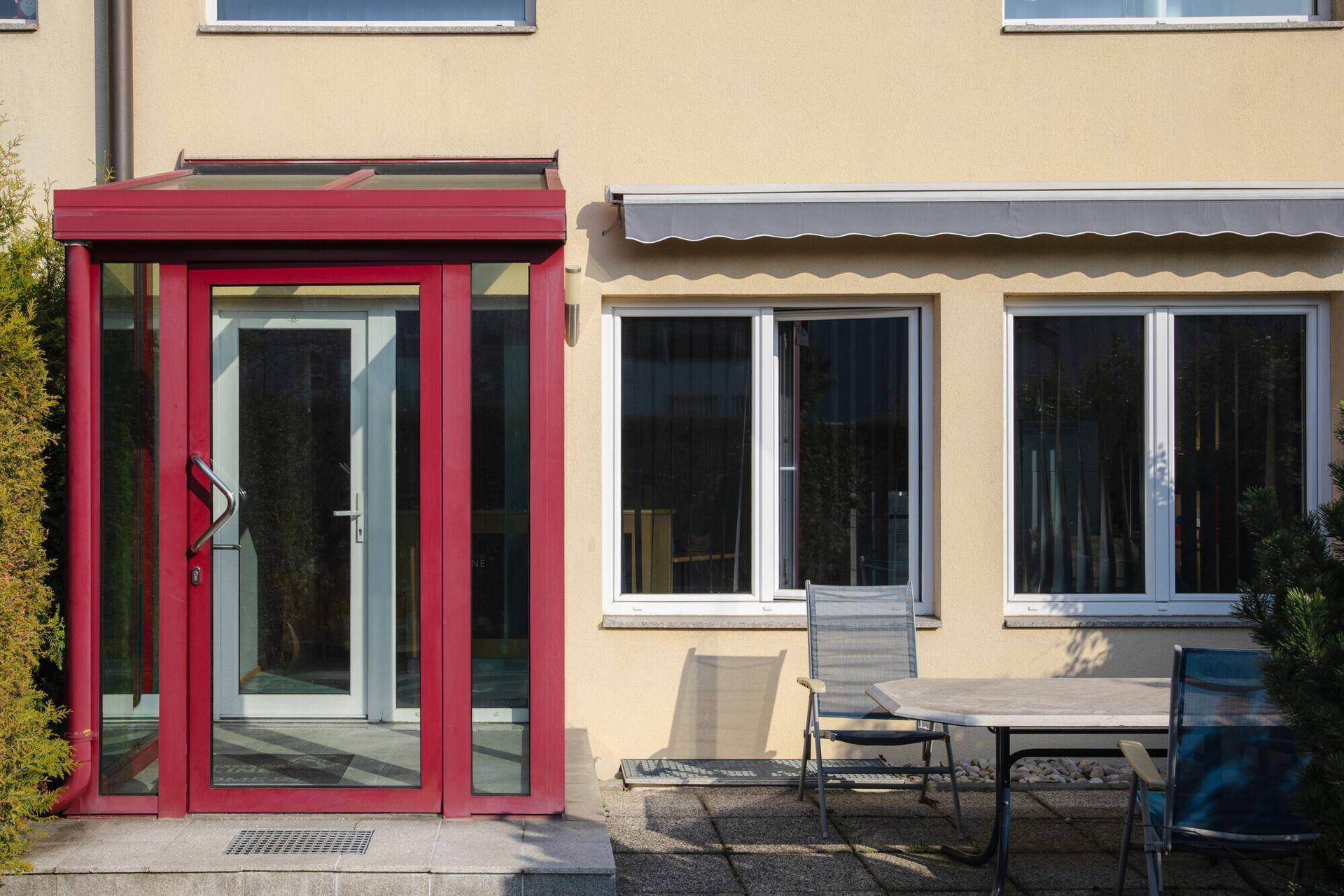 Eingangsbereich mit rotem Vordach und kleiner Terrasse mit Gartenmöbeln, einladend und gemütlich.