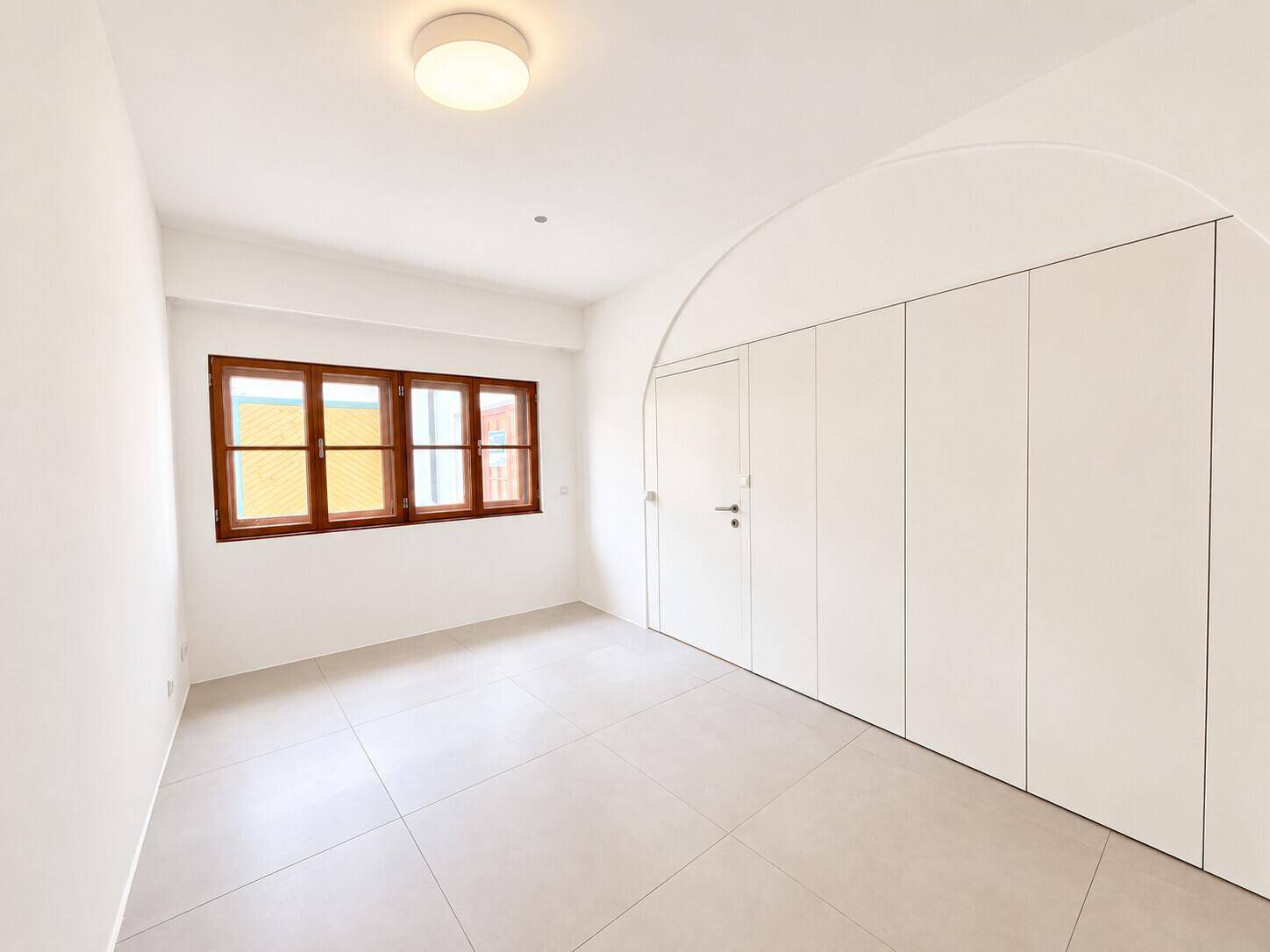 Helles Schlafzimmer mit Fliesenboden, großen Holzfenstern und praktischen Einbauschränken.