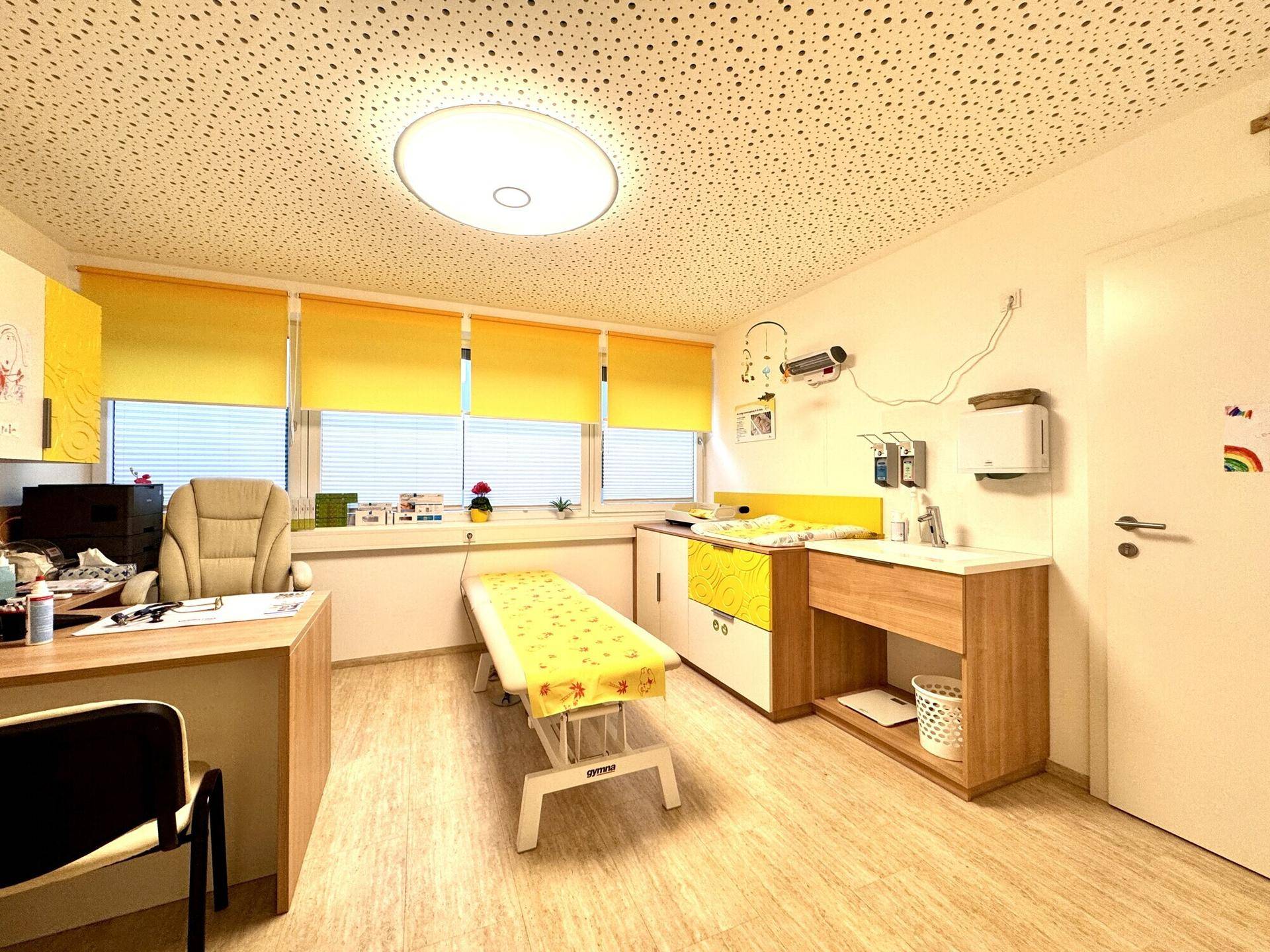 Helles Behandlungszimmer mit Untersuchungsliege, Schreibtisch und gelben Rollos an den Fenstern.