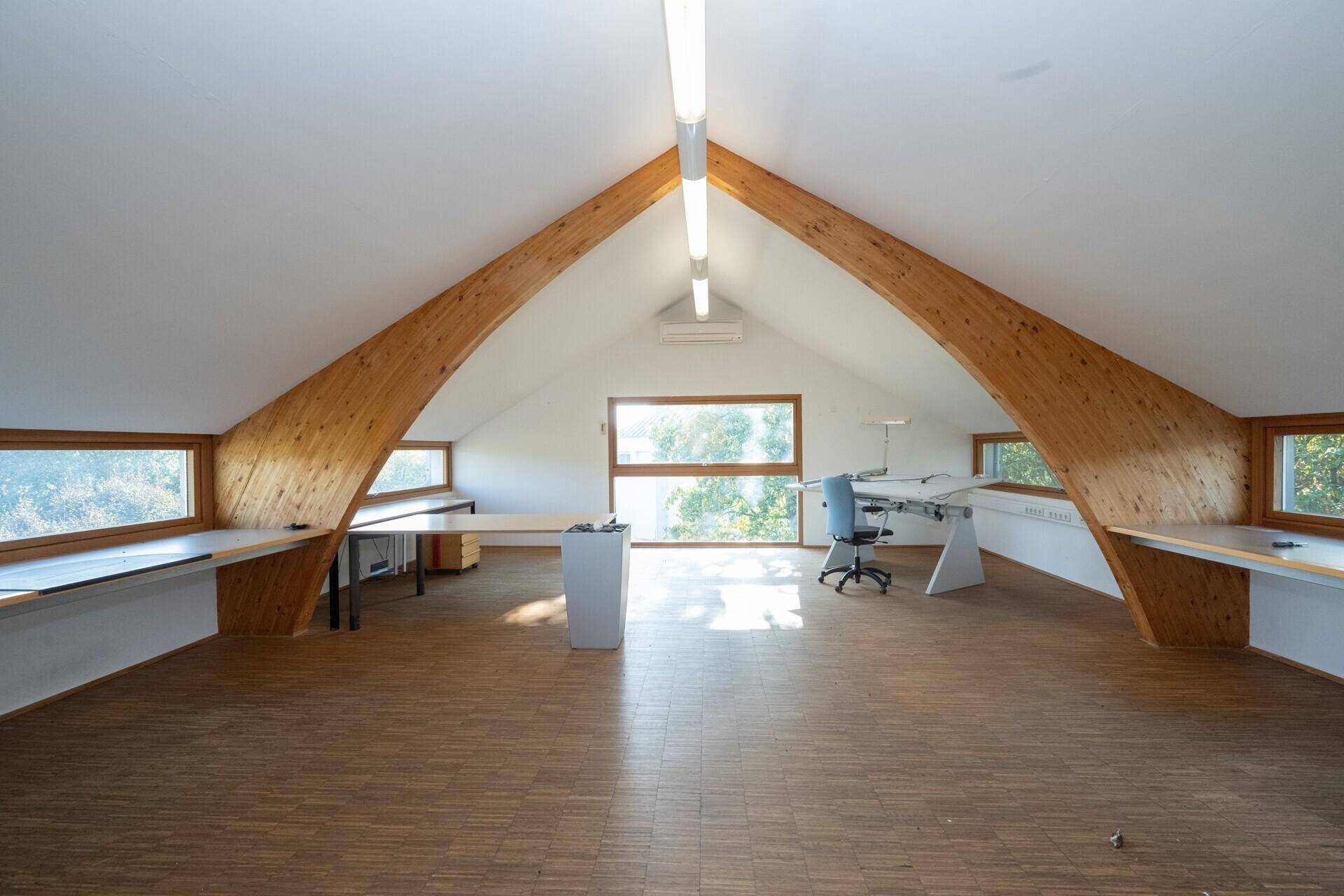 Helles Dachgeschossbüro mit Holzbalken, schrägen Wänden und weitem Ausblick durch die Fenster.