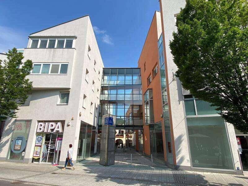 Stadtansicht mit modernen Gebäuden, Geschäften im Erdgeschoss und einer markanten Glasbrücke.