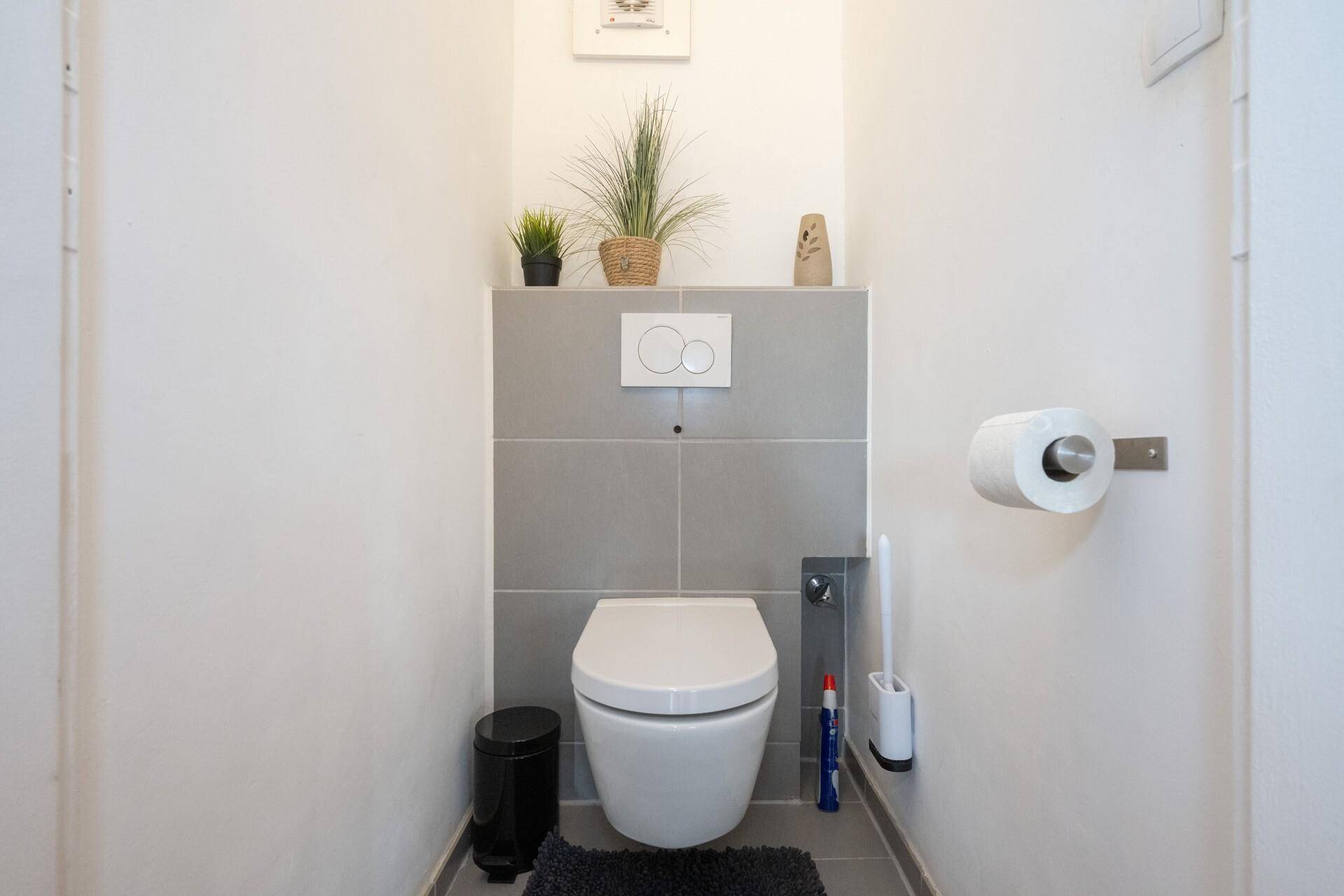 Separates WC mit wandhängender Toilette und grauen Fliesen im Hintergrund.