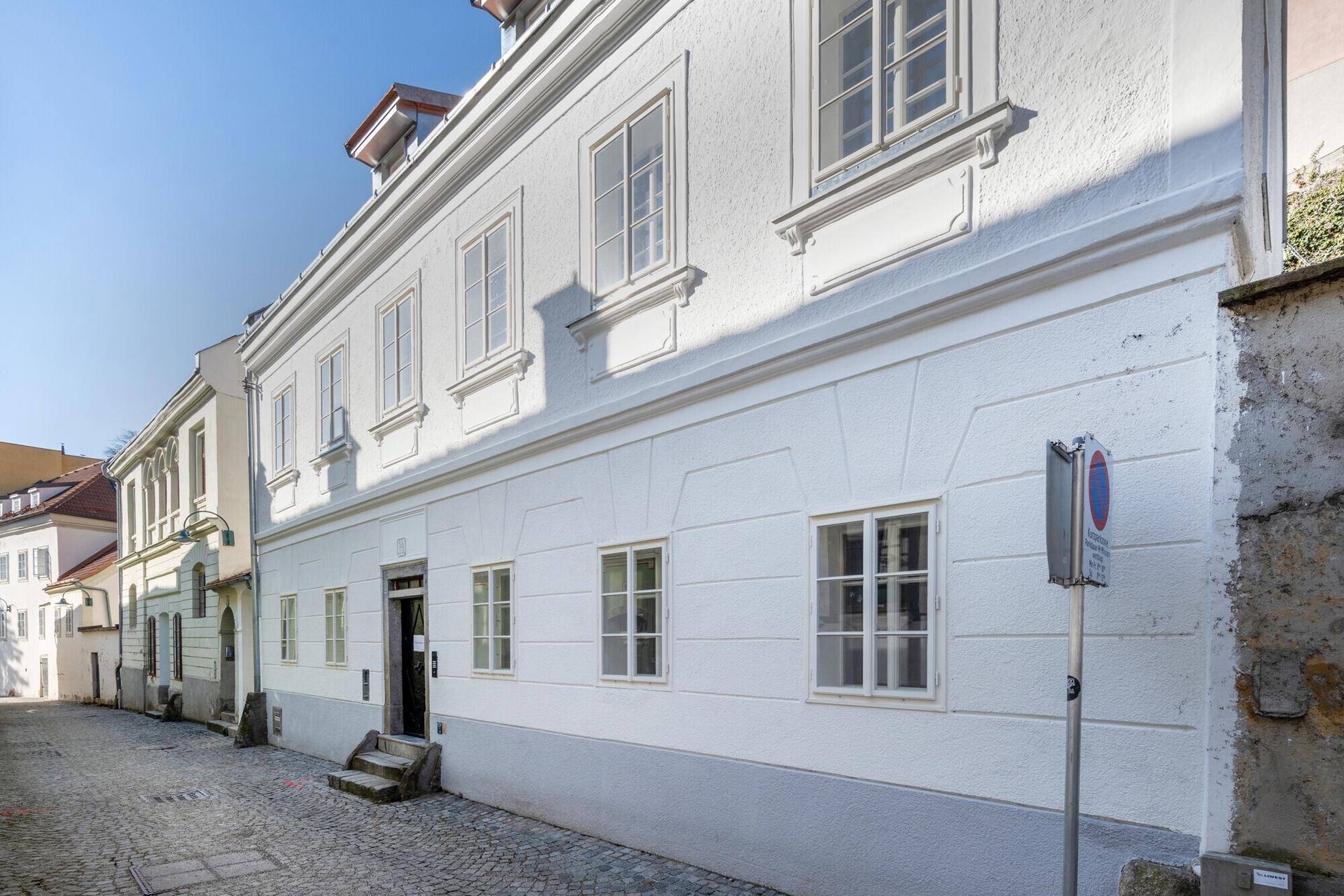 Historisches Gebäude mit weißer Fassade und vielen Fenstern an einer gepflasterten Gasse.