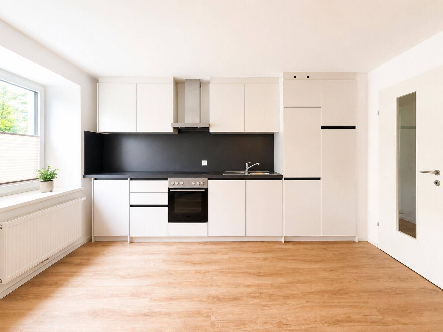 Moderne Küche mit weißen Schränken, schwarzer Arbeitsplatte und Holzfußboden, hell durch ein Fenster beleuchtet.
