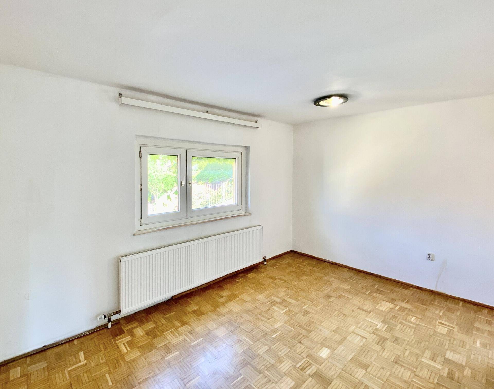 Helles, leeres Zimmer mit Parkettboden, Heizkörper und zwei Fenstern.