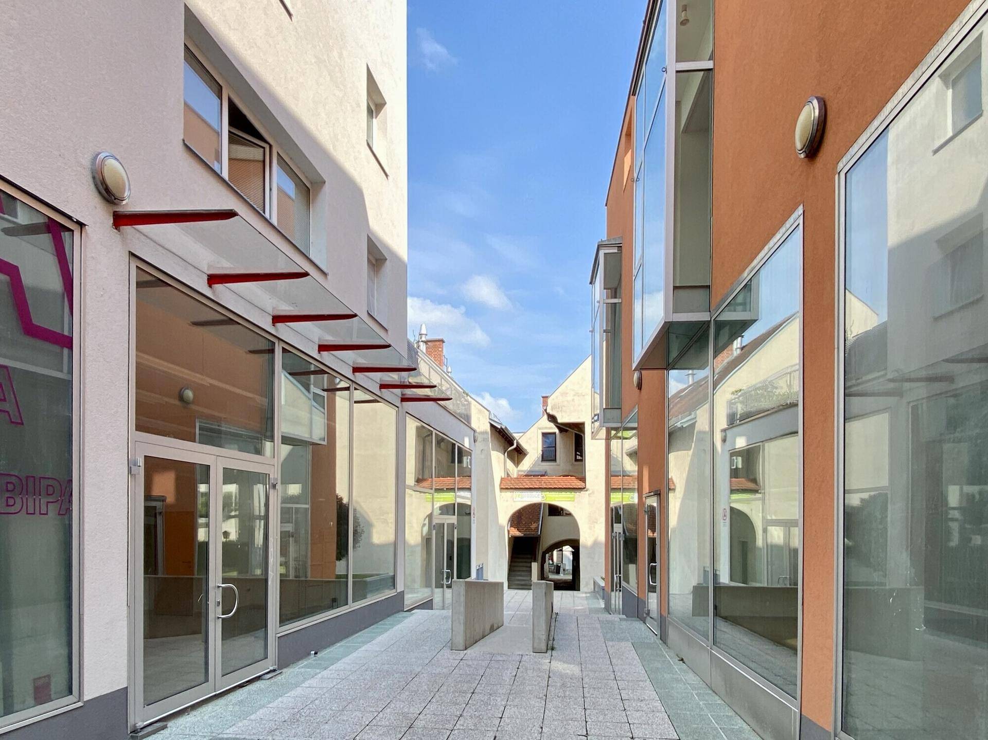 Moderne Gebäudefassaden mit großen Glasflächen säumen eine gepflasterte Gasse unter blauem Himmel.