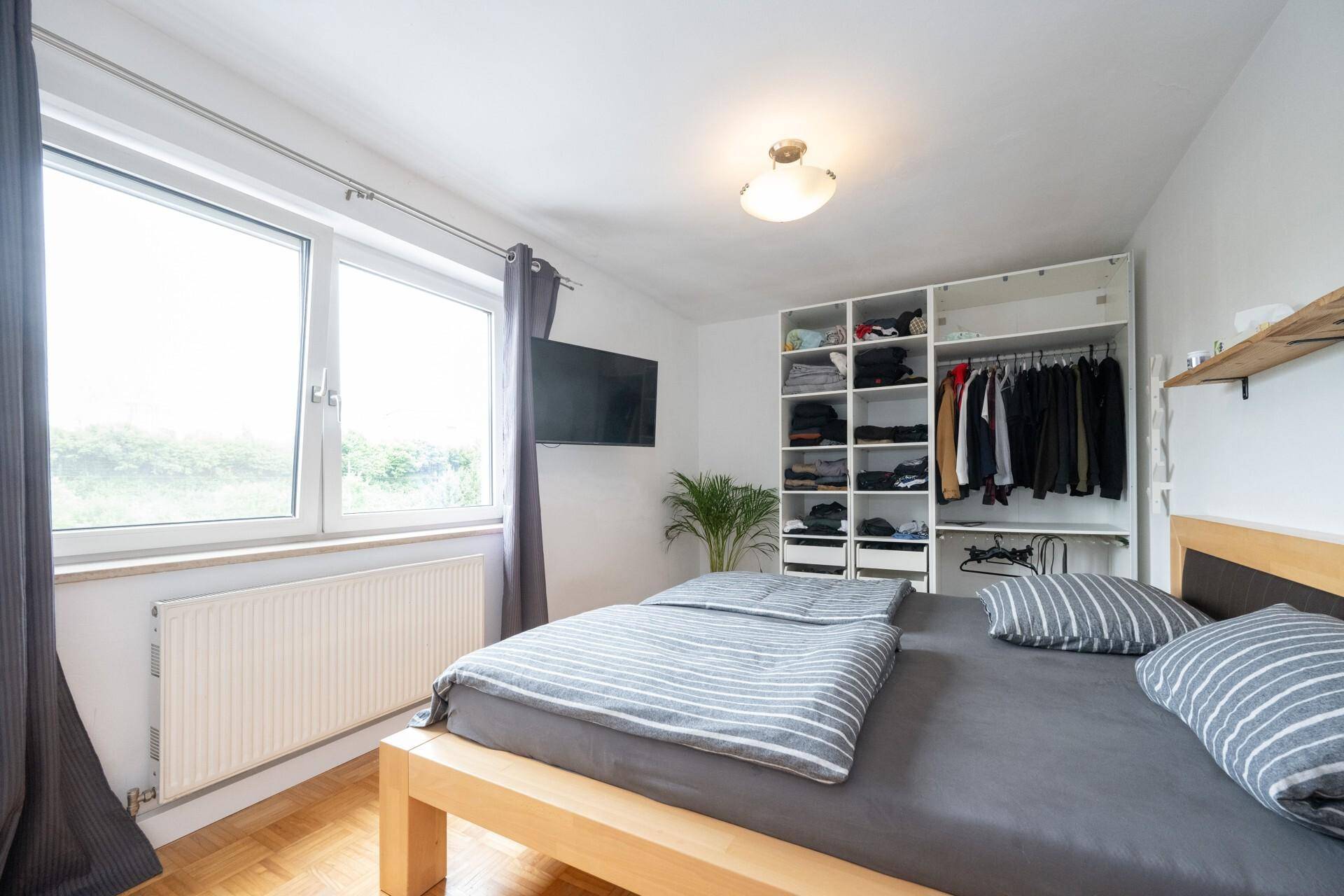 Schlafzimmer mit Holzbett, offenem Kleiderschrank und großem Fenster für viel Licht.