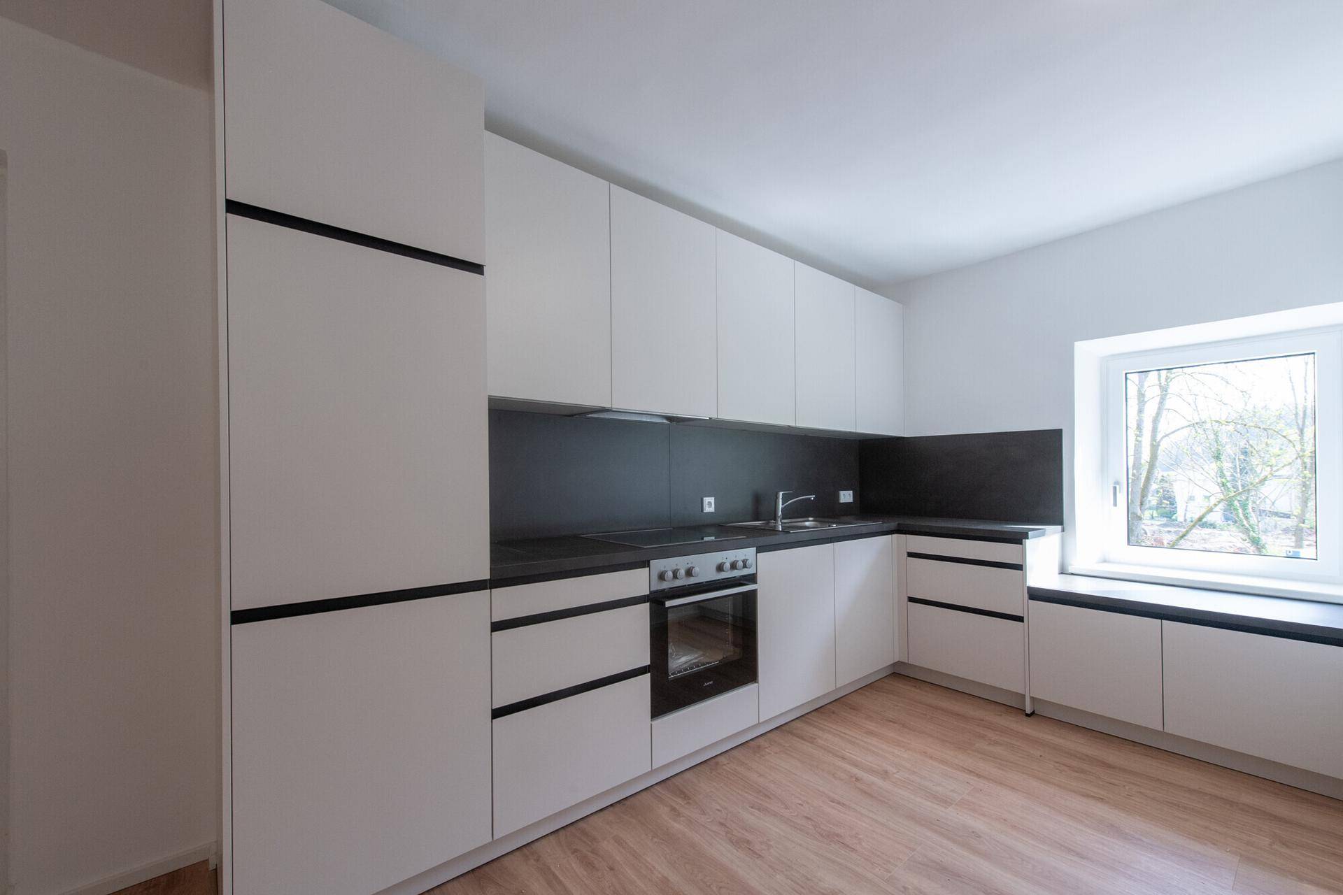 Moderne Einbauküche mit weißen Schränken, dunkler Arbeitsplatte und Fenster mit Blick ins Grüne.