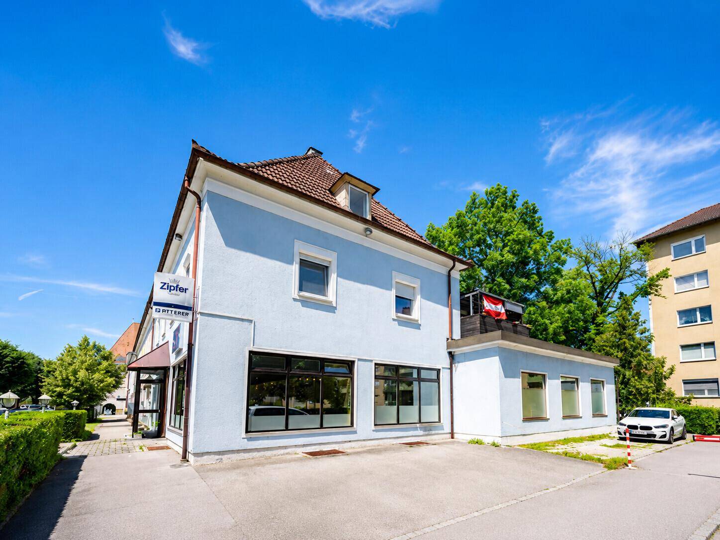 Außenansicht des Gebäudes mit blauer Fassade, großen Fenstern und einem kleinen Parkplatz davor.