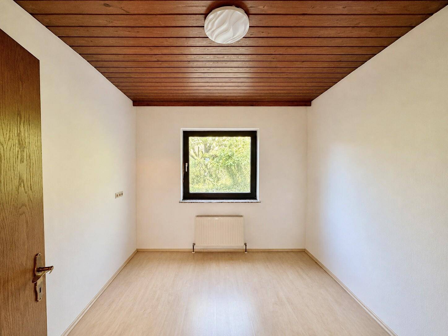 Helles Zimmer mit Holzdecke, Laminatboden und Fenster mit Blick ins Grüne.