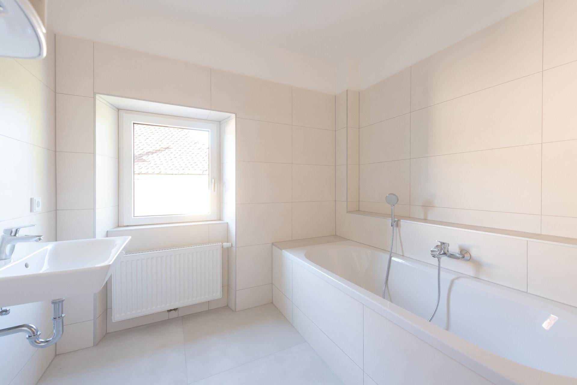 Helles Badezimmer mit weißen Fliesen, Badewanne, Waschbecken und natürlichem Lichteinfall.