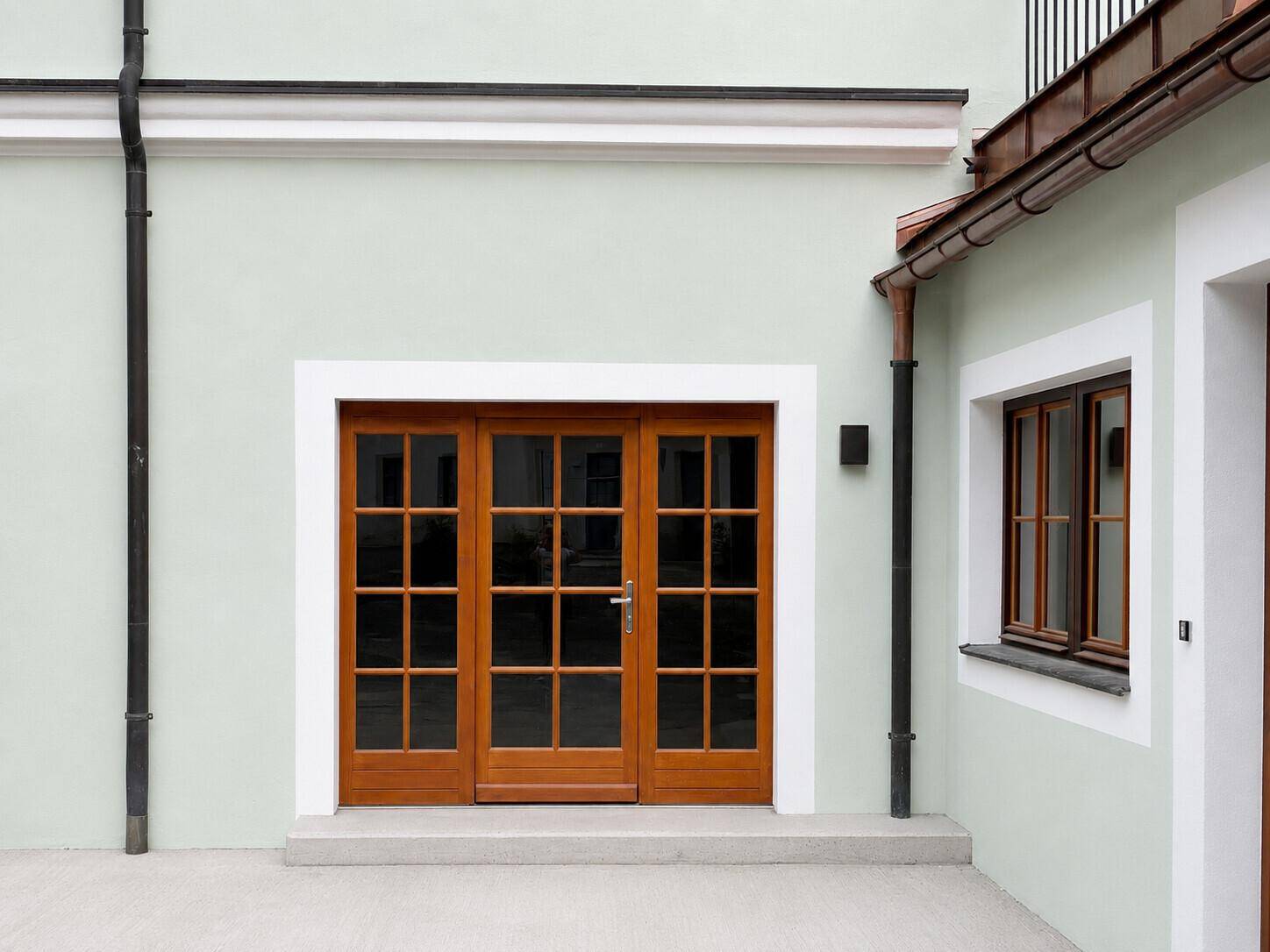Gebäudefassade mit hellgrünem Putz, braunen Holzfenstern und einer passenden Holztür.