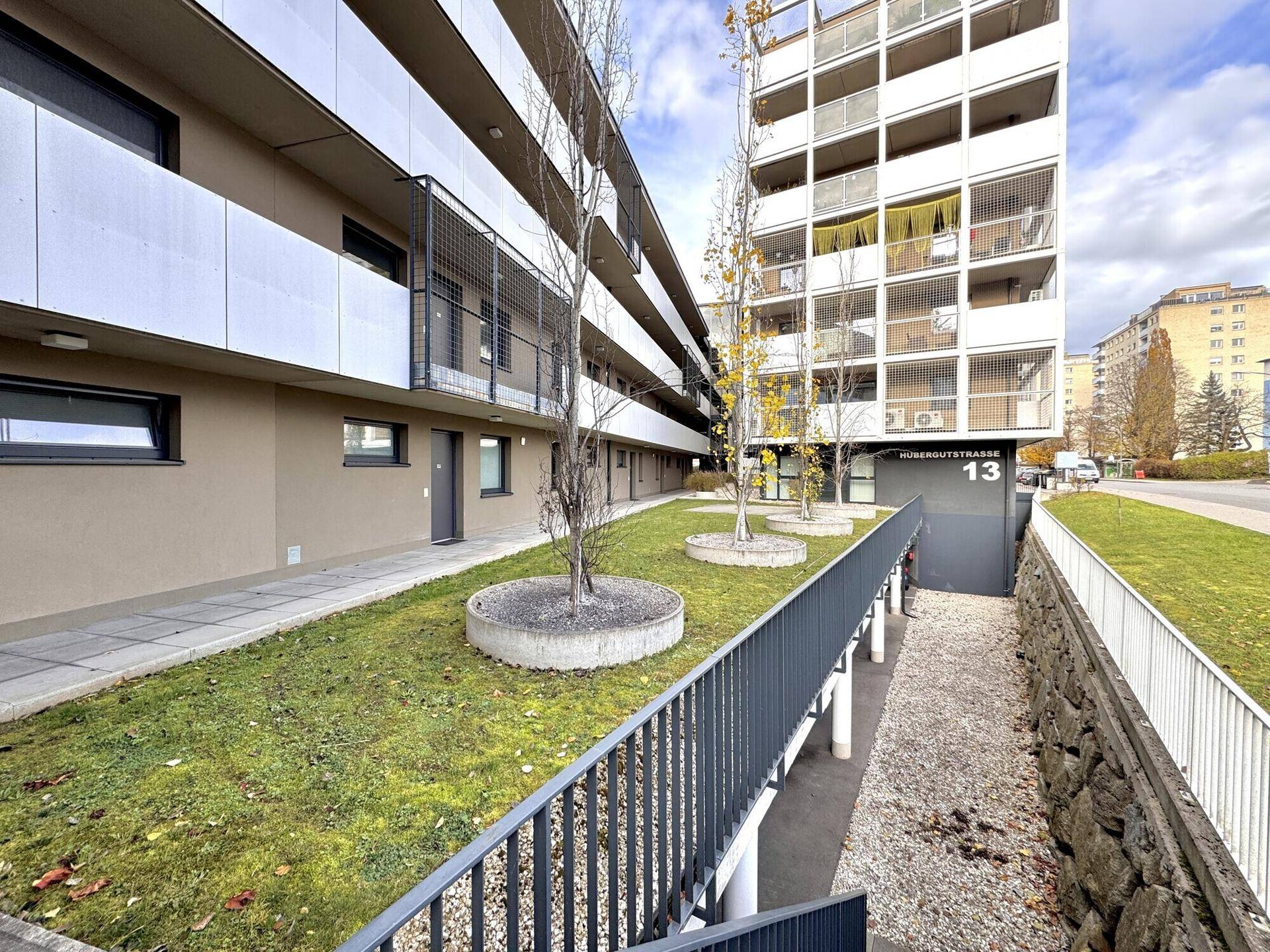 Außenansicht eines modernen Gebäudes mit Grünflächen, einem Gehweg und einer Rampe für barrierefreien Zugang.