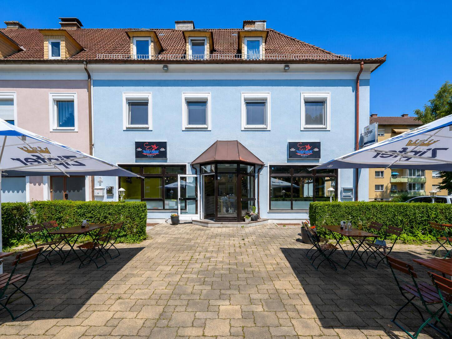Frontansicht des Gebäudes mit Eingangsbereich und einer gepflasterten Terrasse mit Tischen und Sonnenschirmen.