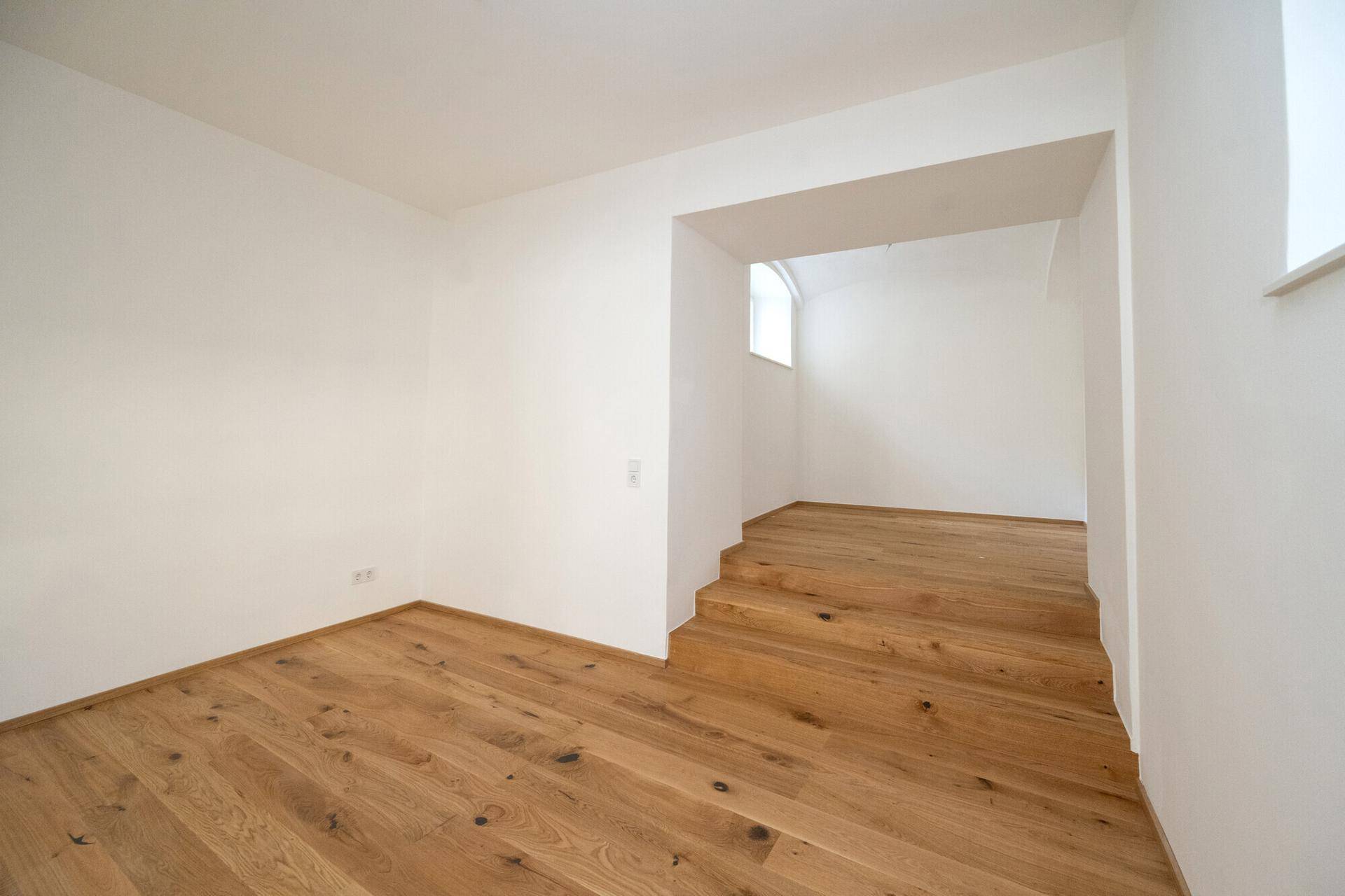 Geräumiger, heller Raum mit Holzboden und einer Treppe, die in einen weiteren Bereich führt.