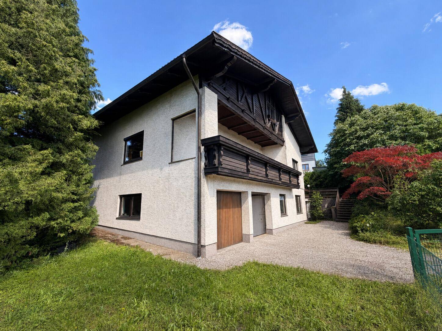 Seitenansicht des Hauses mit Holzbalkon, Garagentor und Außentreppe in ländlicher Umgebung.