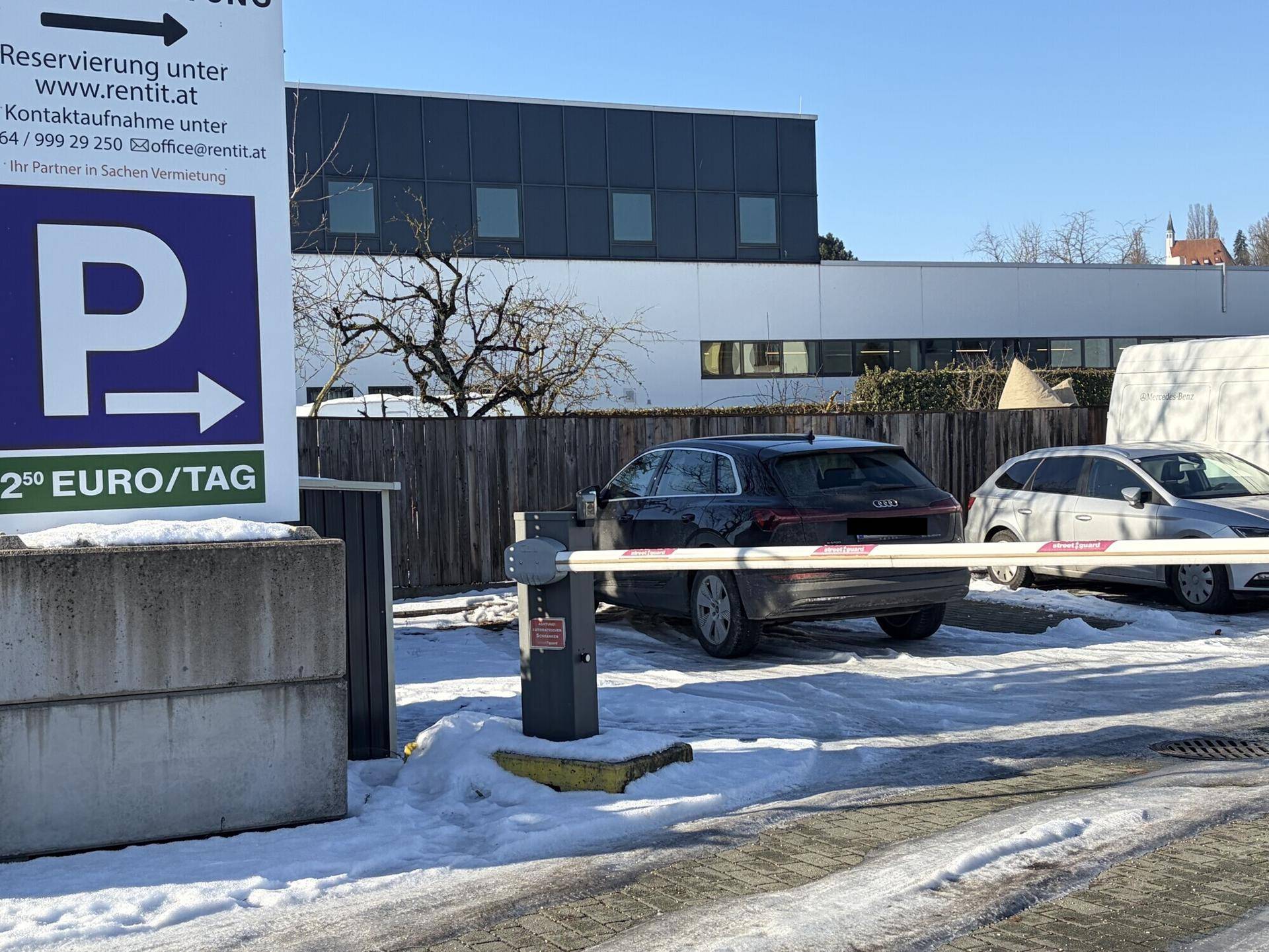 Ein Auto fährt durch die geöffnete Schranke eines Parkplatzes mit Hinweisschildern im Hintergrund.