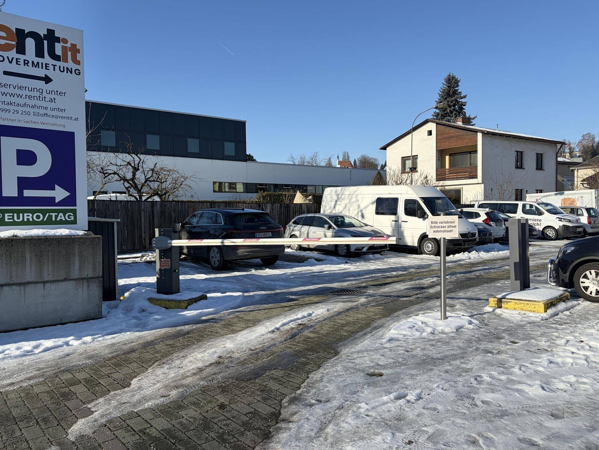 Überblick über den Eingangsbereich eines Parkplatzes mit mehreren geparkten Fahrzeugen und Schnee auf dem Boden.