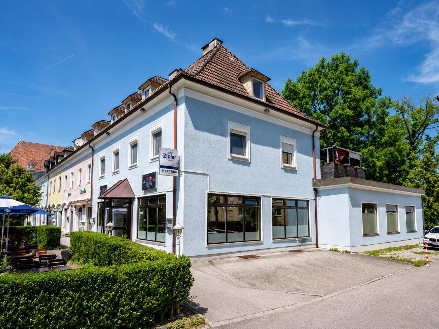 Gebäudeansicht mit blauer Fassade, Dachgauben und einer einladenden Außenterrasse mit Sonnenschirmen.