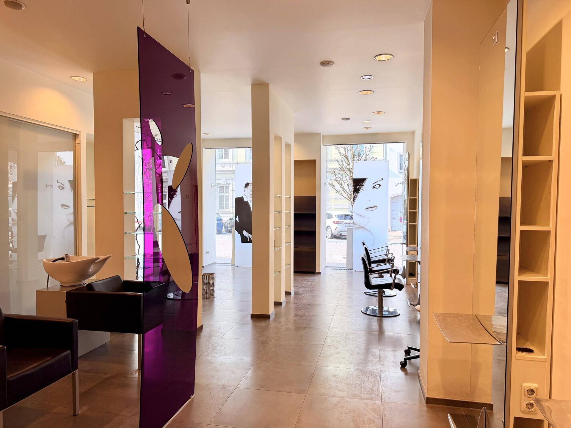 Heller Friseursalon mit großen Schaufenstern, lila Dekorationselementen und modernen Frisierplätzen.