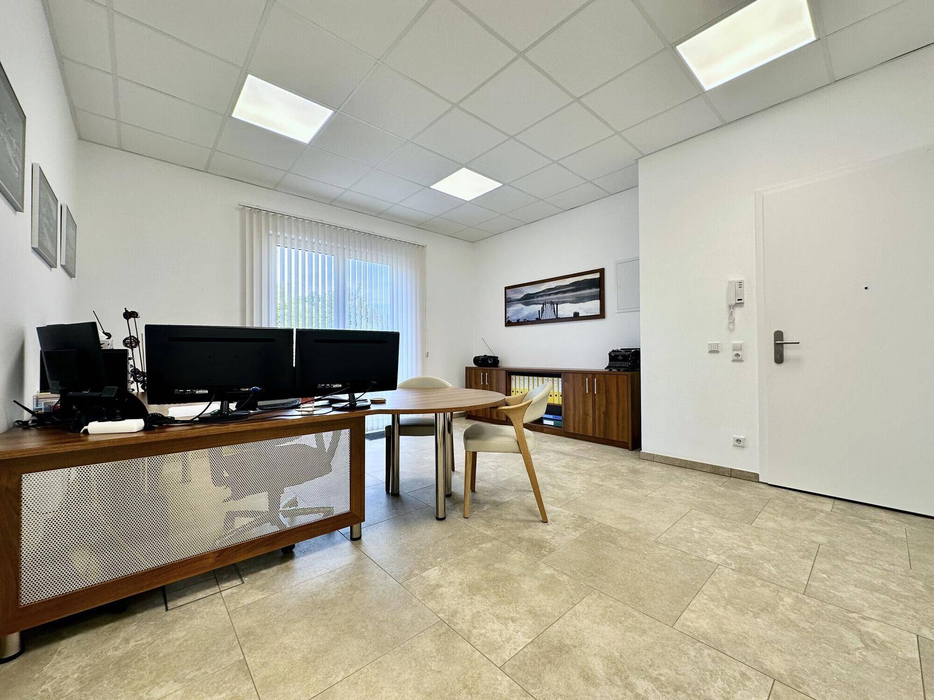 Heller Bürobereich mit Fliesenboden, Holzschreibtisch und Zugang zur Eingangstür.