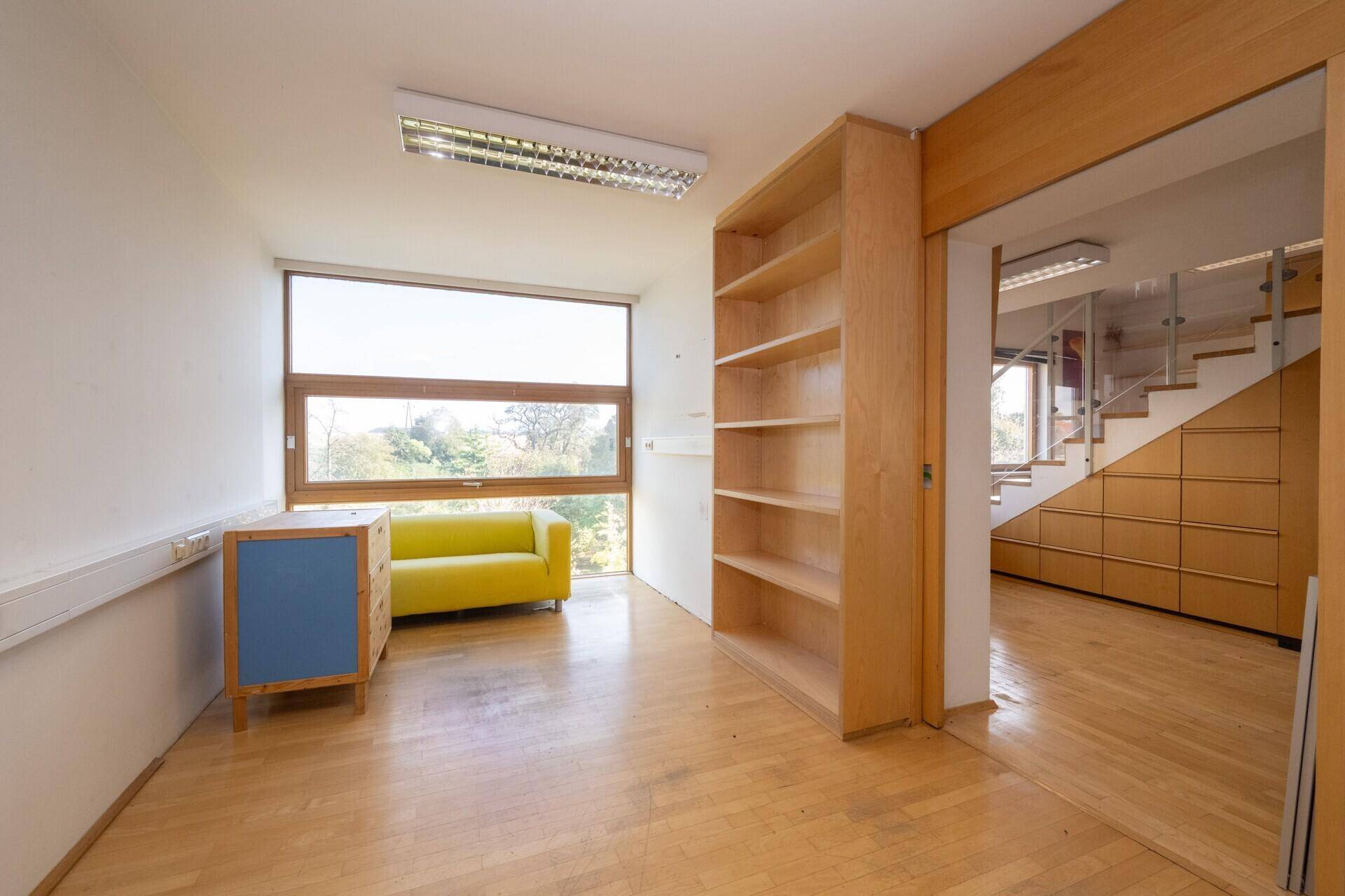 Helles Büro mit Holzparkett, großem Fenster und einem auffälligen gelben Sofa.