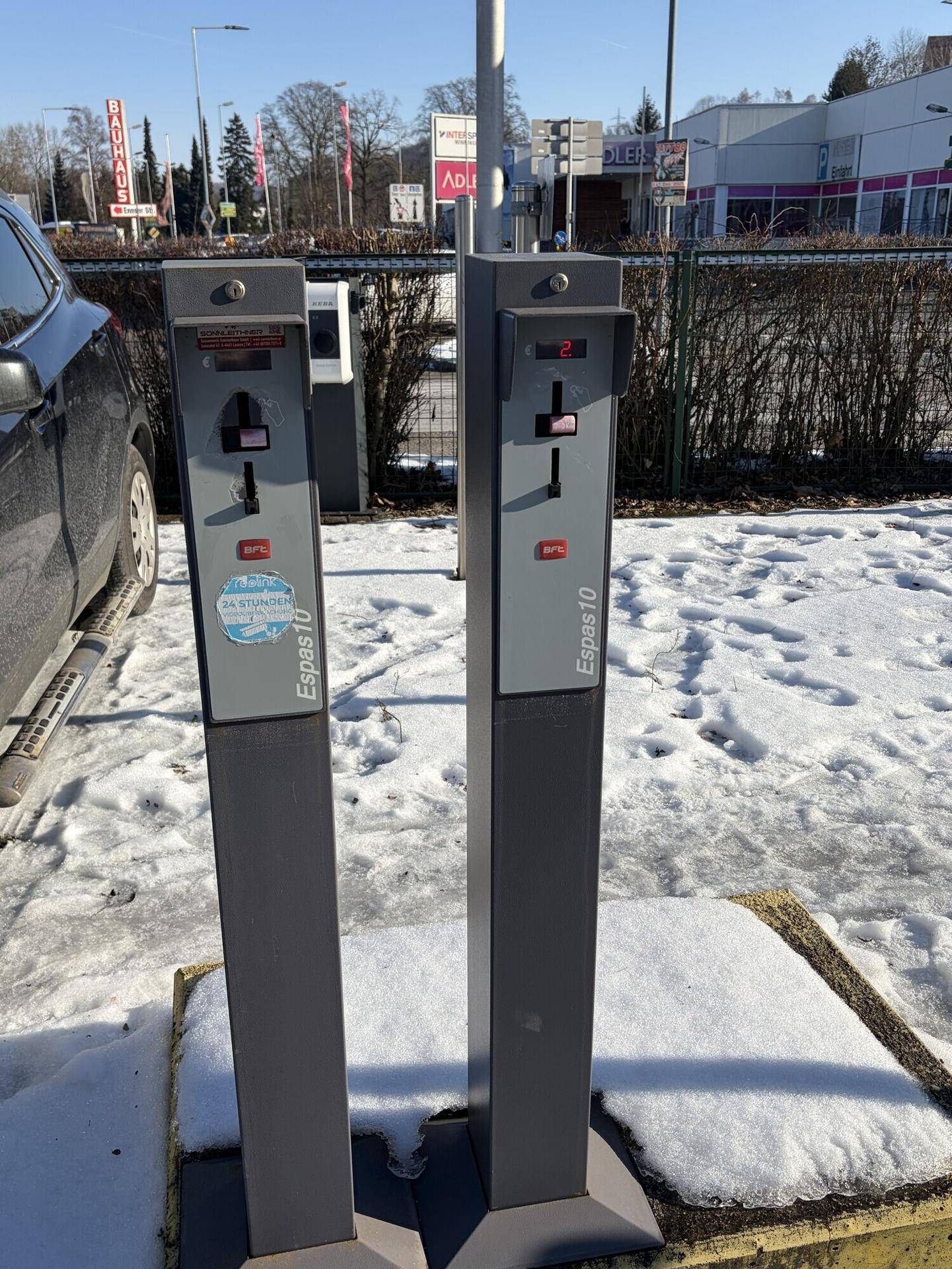 Zwei Parkautomaten stehen auf einem verschneiten Parkplatz mit einem geparkten Fahrzeug im Vordergrund.