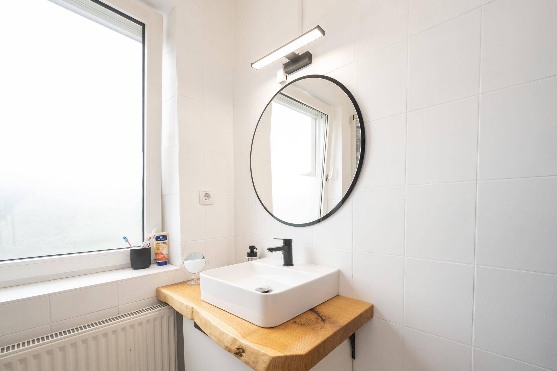 Modernes Badezimmer mit Aufsatzwaschbecken, rundem Spiegel und weiß gefliesten Wänden.