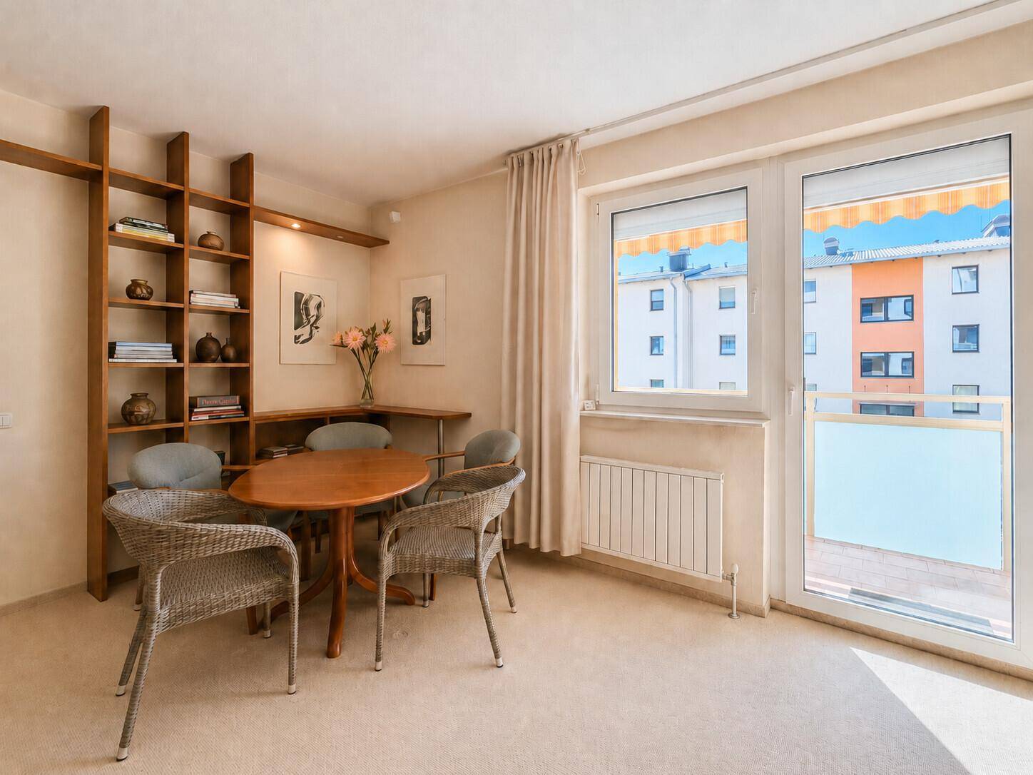 Essbereich mit Teppichboden, Holzregal und Zugang zu einem Balkon mit Blick auf die Umgebung.
