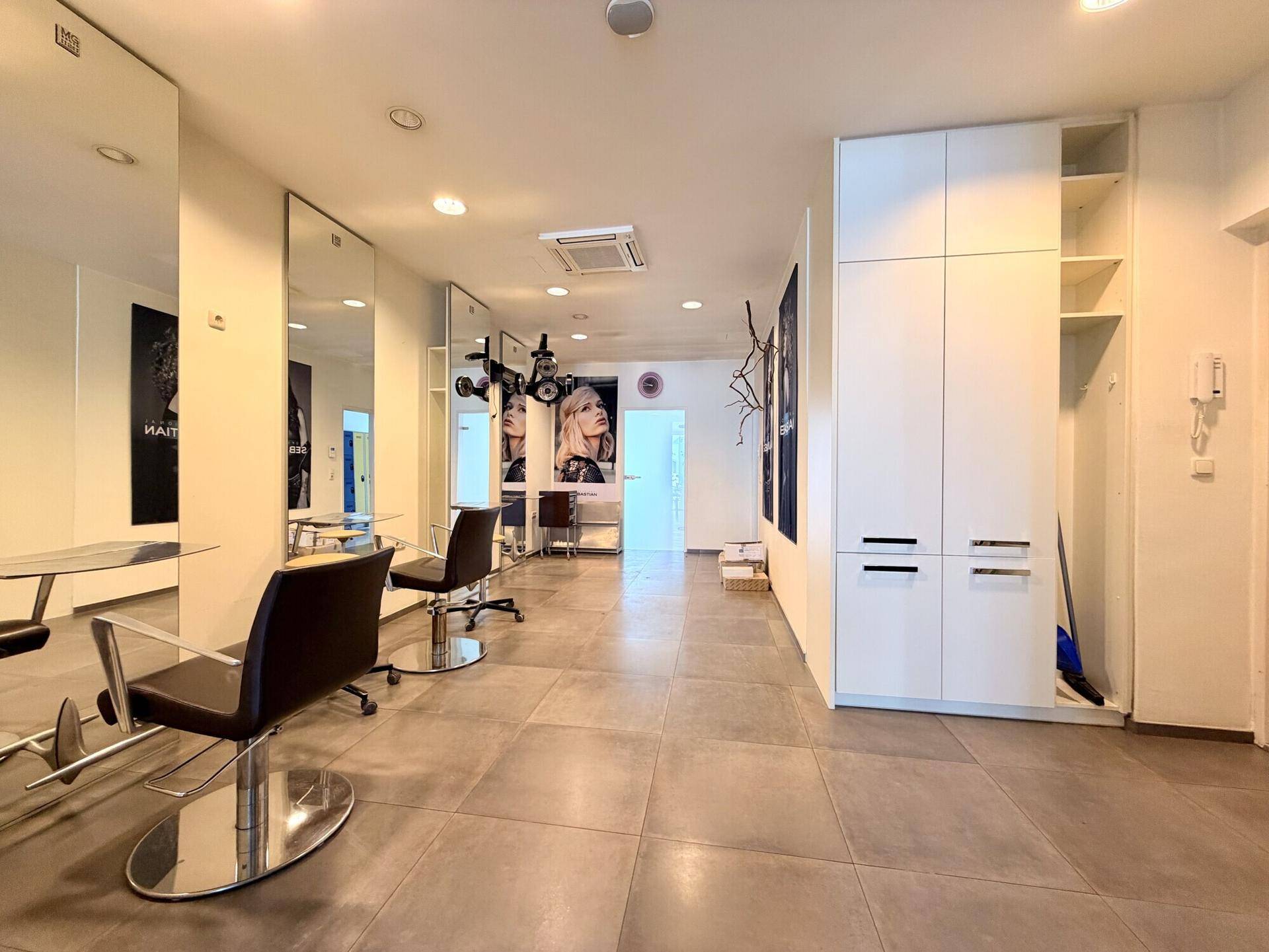 Geräumiger Friseursalon mit weißen Einbauschränken, großen Spiegeln und Fliesenboden.