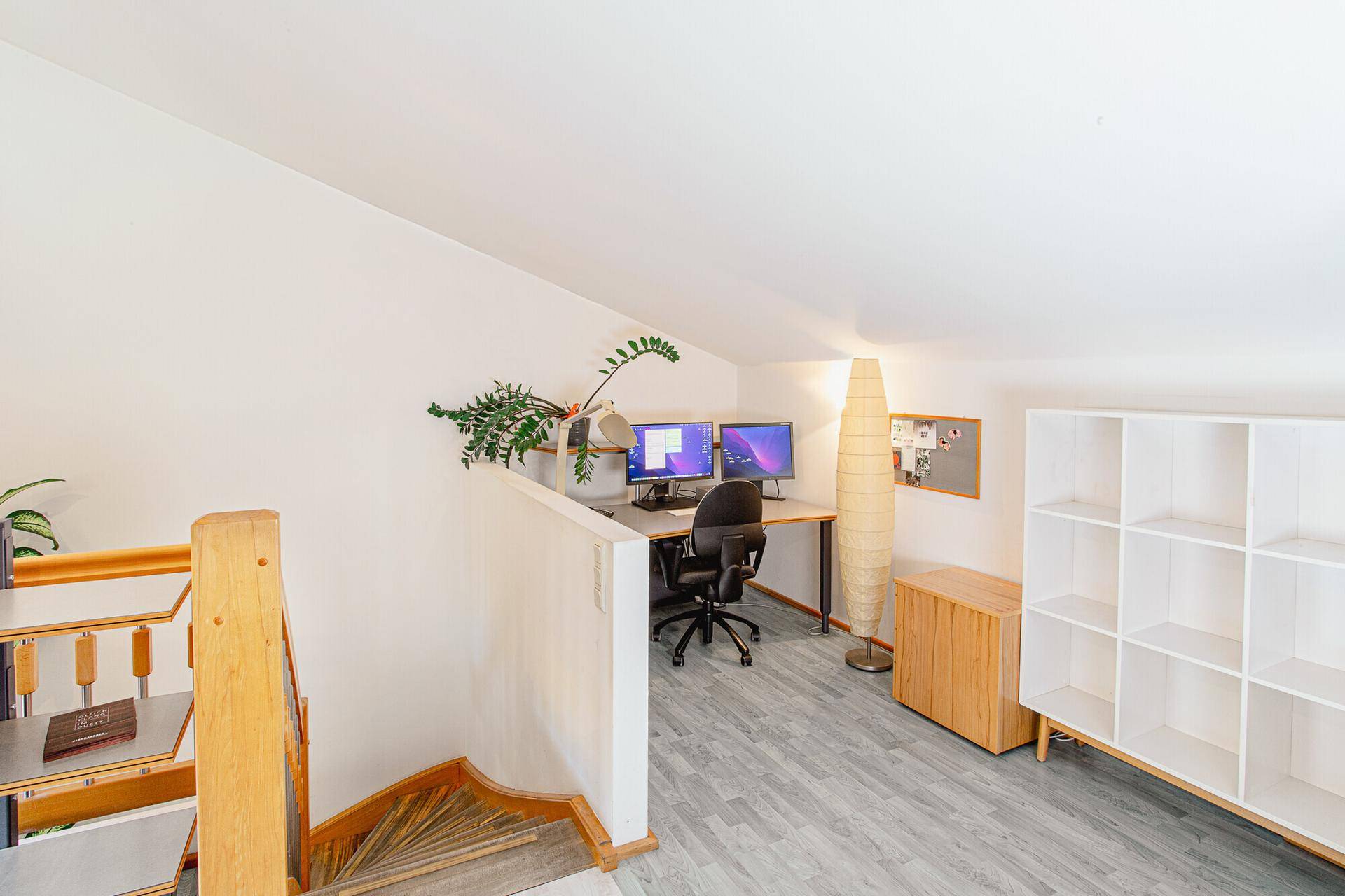 Helles Büro im Obergeschoss mit Dachschräge, Schreibtisch, Computermonitoren und einem Bücherregal.