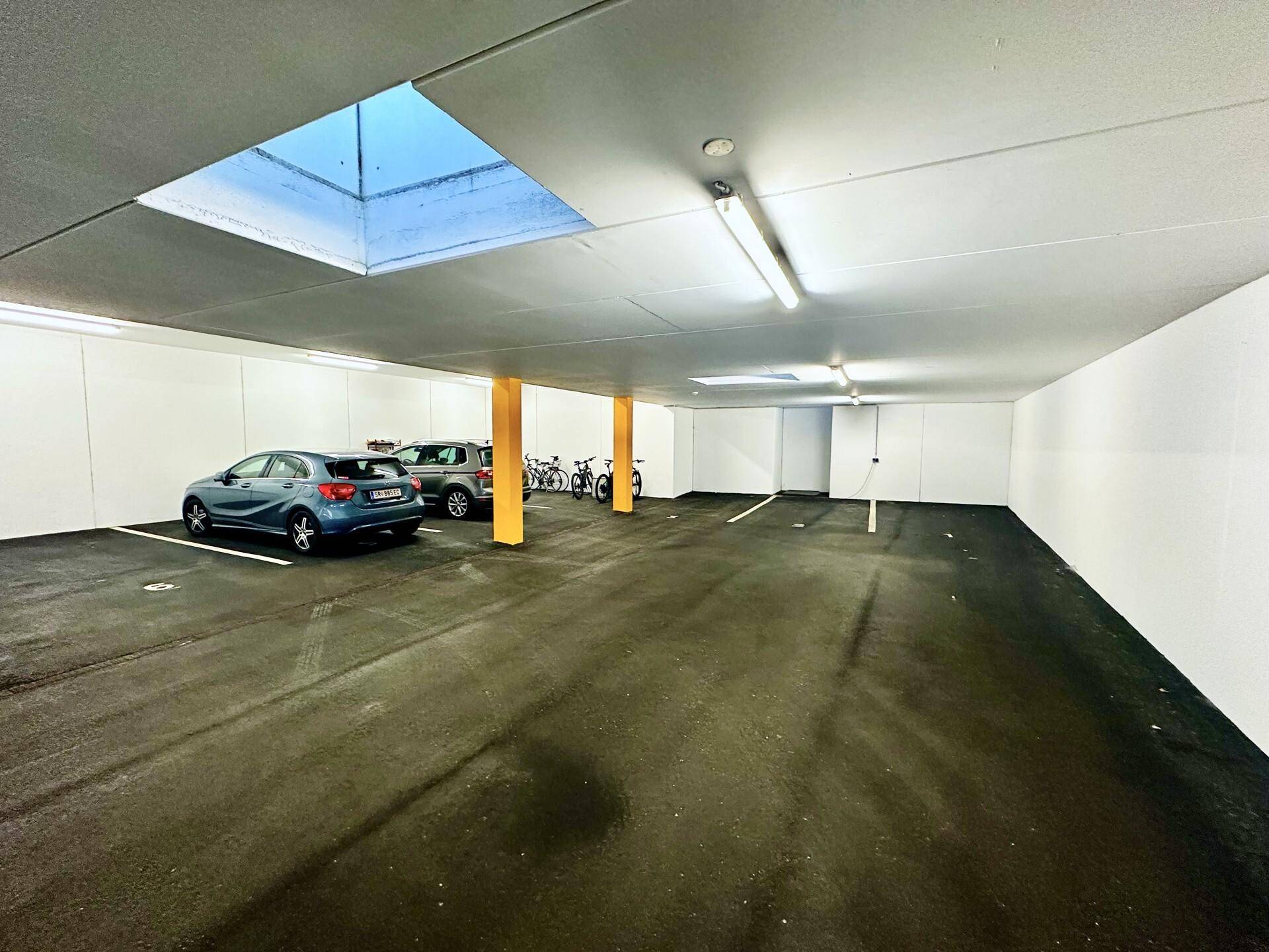 Tiefgarage mit geparkten Autos und Fahrrädern, beleuchtet durch ein Oberlicht.